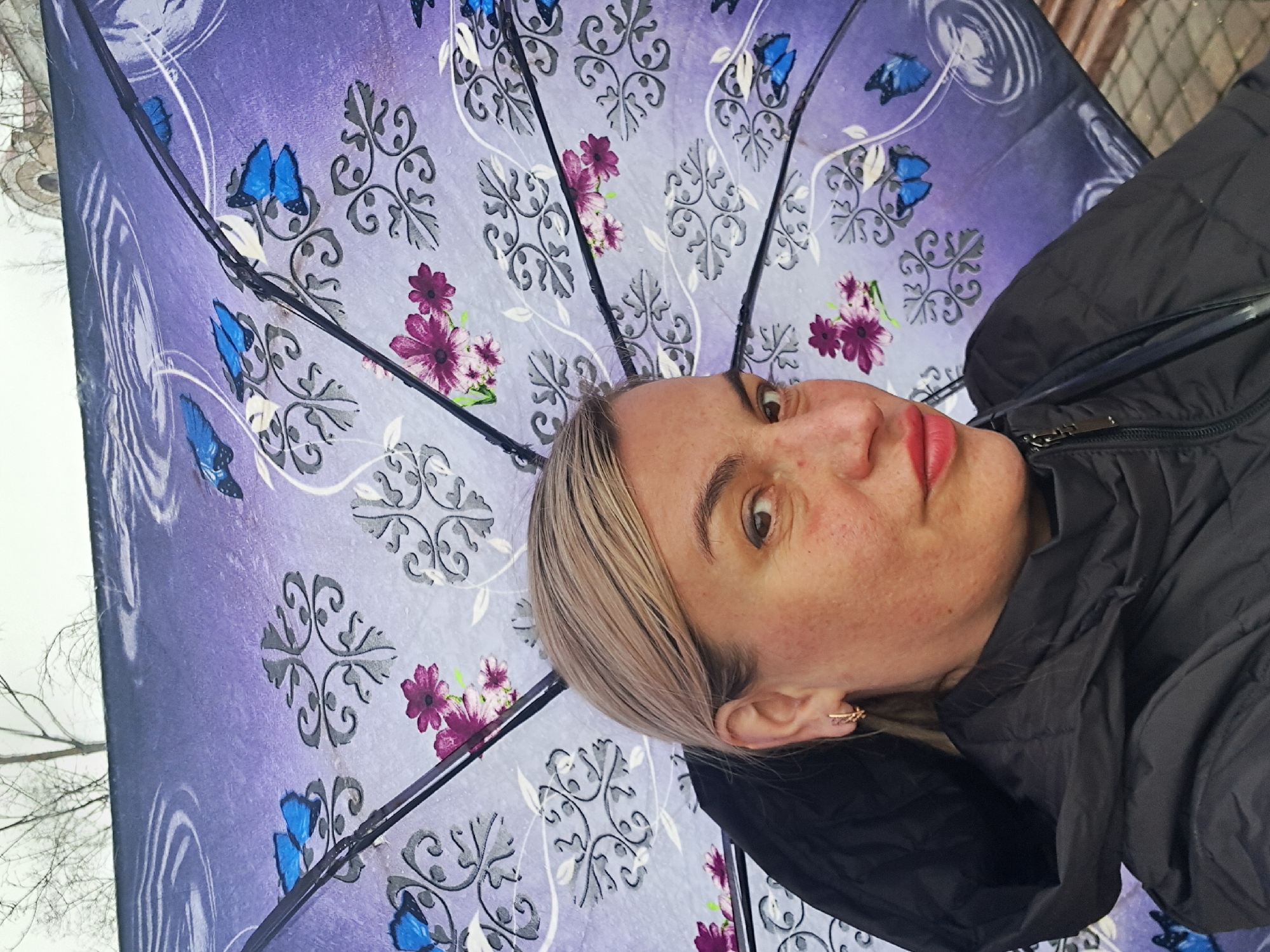 A woman stands under a purple umbrella with a pattern of flowers, butterflies, and swirling designs. She is wearing a black hooded jacket. The background shows trees with bare branches and a possibly gray or overcast sky, suggesting an outdoor setting. The photograph appears to have been taken at a slight angle. The city of Elektrostal, Russia is not evident.
