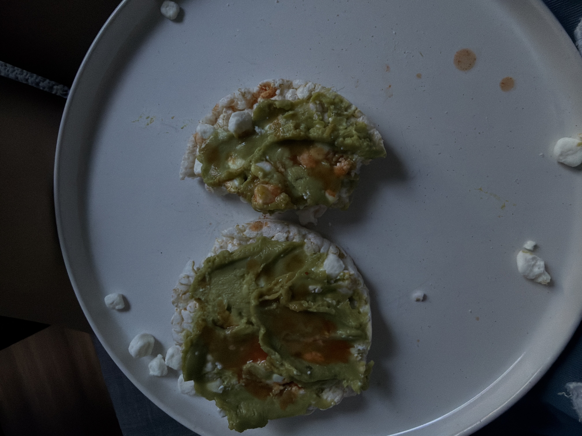 A top-down, indoor shot depicts two rice cakes on a white, round plate. Each rice cake is topped with a layer of green mashed avocado and a reddish-orange sauce. Small white cheese pieces are scattered around the rice cakes on the plate. Some small brown stains and crumbs are also visible on the plate. The left edge of the plate is near a wooden surface, while a blue fabric is seen on the right. The image does not provide any information to place the scene in New York City or the United States of America.