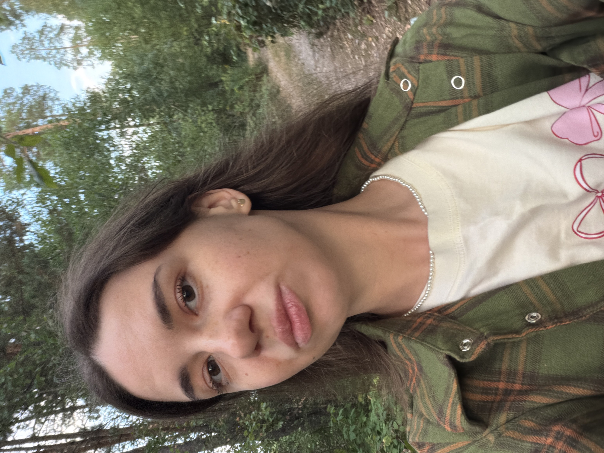 The image is a selfie taken outdoors, likely in a wooded area. The subject, a young woman with dark hair, is looking at the camera with her mouth slightly puckered. She is wearing a green and orange plaid shirt over a cream-colored t-shirt with a pink bow graphic. She has a pearl necklace on. The background consists of trees and a path, suggesting a natural, possibly rural, environment. The overall lighting indicates it is daytime. The provided hints focus on different parts of the subject's face, clothing, and the background, allowing for closer examination of these elements. There is no information within the image that explicitly places it in the city of Kmytiv, Ukraine.