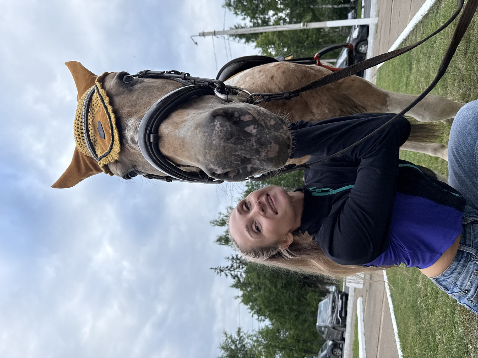 The image depicts a close-up of a woman and a horse in Chaikovsky, Russia. The woman, with blonde hair tied in a ponytail, is wearing a black jacket over a blue top and jeans. She is smiling and leaning towards the horse. The horse, a light brown color, is wearing a brown ear net with a decorative trim and a bridle. The background shows a cloudy sky, trees, grass, and a street with cars. The scene appears to be outdoors, possibly in a park or equestrian facility.
