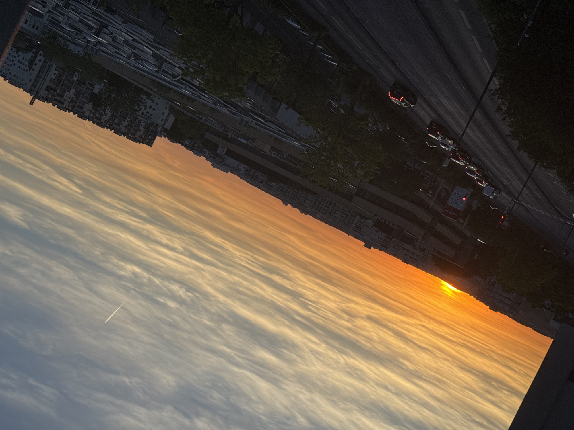 A wide-angle, high-angle, tilted shot captures a sunset over an urban landscape. The sky is a gradient of warm oranges and yellows near the horizon, transitioning to pale blues and whites with wispy clouds higher up. A faint contrail from an airplane is visible in the upper right quadrant. In the foreground, a street curves away from the viewer, lined with trees and buildings. Several cars are visible on the street, some with their headlights on, suggesting late evening. A traffic light is red. Beyond the street, a collection of buildings, including apartment blocks and what appears to be commercial or industrial structures, are visible. The overall impression is one of a city at dusk, bathed in the soft light of a setting sun.