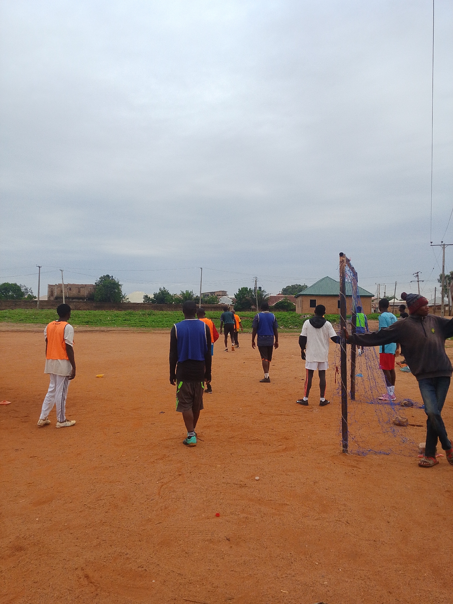 The scene depicts a group of people in an open, sandy area possibly used for sports, as indicated by the net and cones visible. The individuals are wearing casual athletic attire. The sky is overcast, suggesting dull lighting. Buildings and vegetation are visible in the background. Location: Unknown, Unknown.