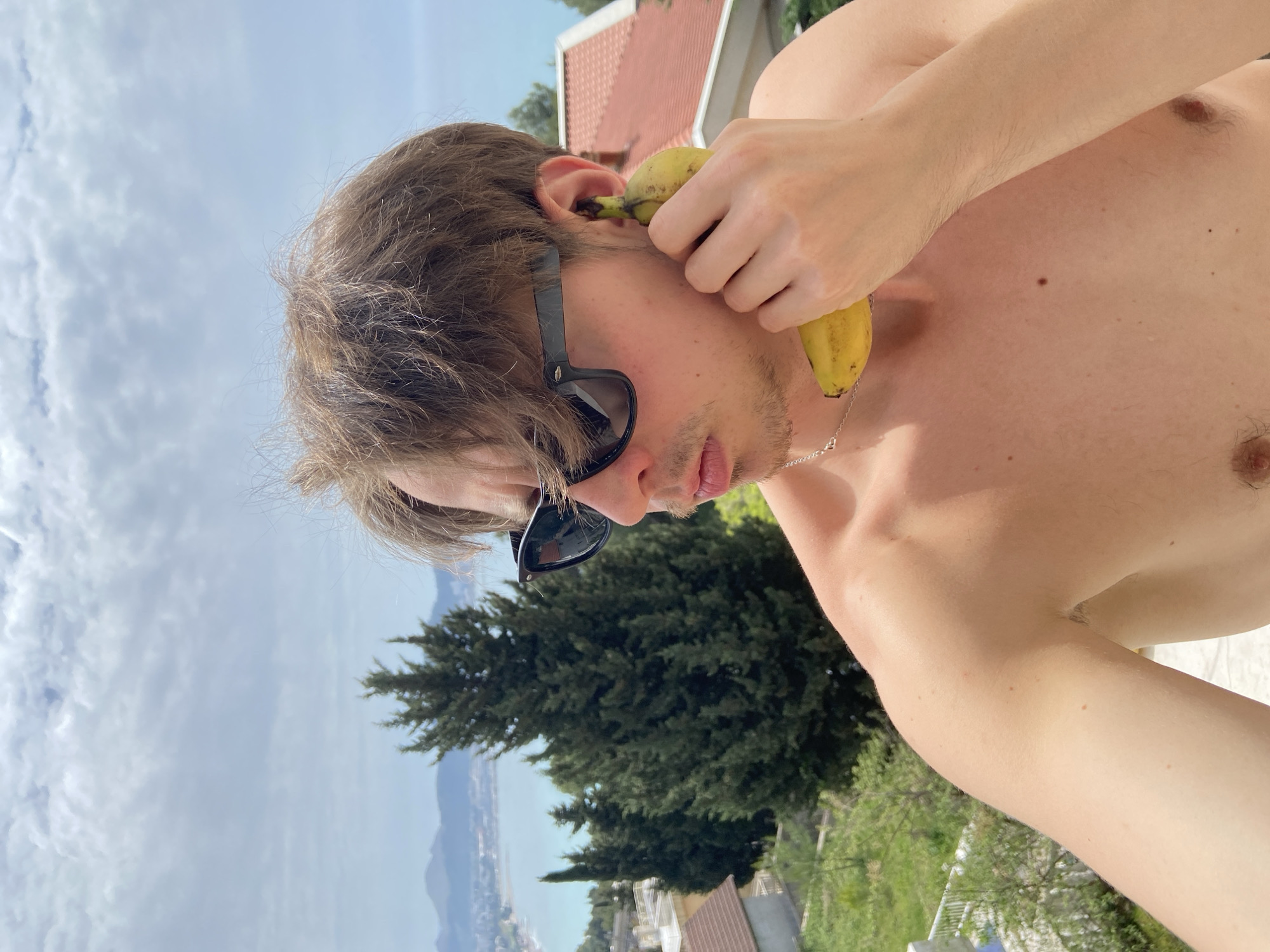 A shirtless man with sunglasses holds a banana to his ear. The background features a cloudy sky, a green tree, and a view of the coast with a body of water. In the far background, there are buildings with orange tile roofs. The location appears to be Šušanj, Montenegro, based on the coastal scenery and building styles.