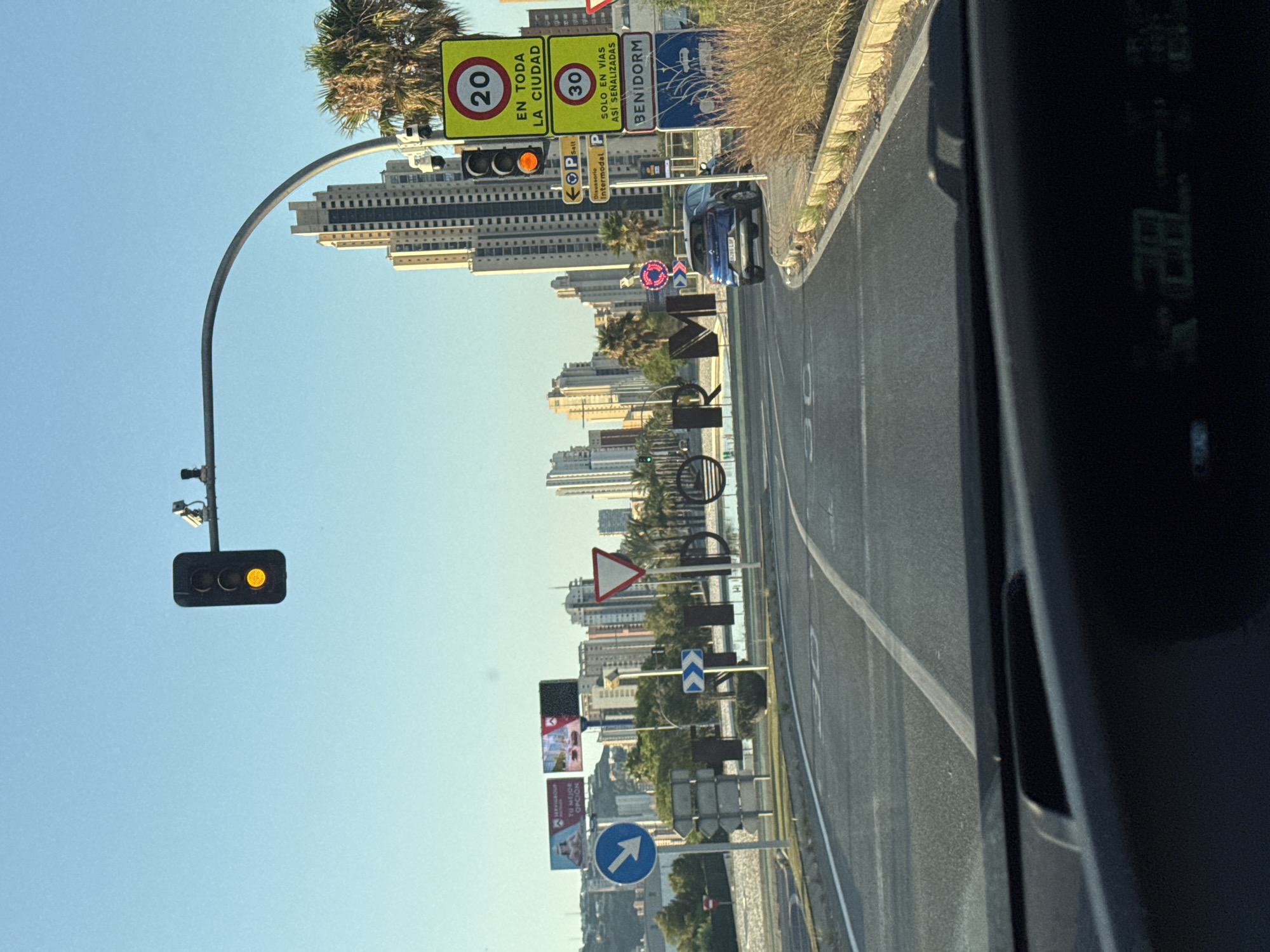 The image captures a street scene in Benidorm, Spain, likely in the early morning or late afternoon due to the low angle of the sun casting long shadows. In the foreground, the dashboard of a vehicle is visible, displaying a digital readout, possibly of temperature or speed. The road ahead is asphalt with white lane markings and speed limit indicators (30) painted on the surface.
To the right of the road, a series of traffic and informational signs are mounted on poles. These include a traffic light showing amber, a speed limit sign indicating 20 km/h "EN TODA LA CIUDAD" (in the entire city), another speed limit sign indicating 30 km/h "SOLO EN VIAS ASI SEÑALIZADAS" (only on signposted roads), and a sign for "BENIDORM". There are also parking signs and a directional sign for "Disuasorio Intermodal".
Further down the road, a blue car is parked or driving. In the mid-ground, large black letters spell out "BENIDORM" in a stylized font, forming a prominent landmark. Behind this, a body of water or canal is visible, reflecting the sky. On the left side of the road, a blue circular sign with a white arrow pointing downwards indicates a direction. Several other smaller signs and poles are also present.
The background is dominated by a cluster of tall, modern high-rise buildings characteristic of Benidorm's skyline, interspersed with palm trees. The sky is clear and a pale blue. The overall impression is a well-developed urban environment with a strong presence of tourism infrastructure.