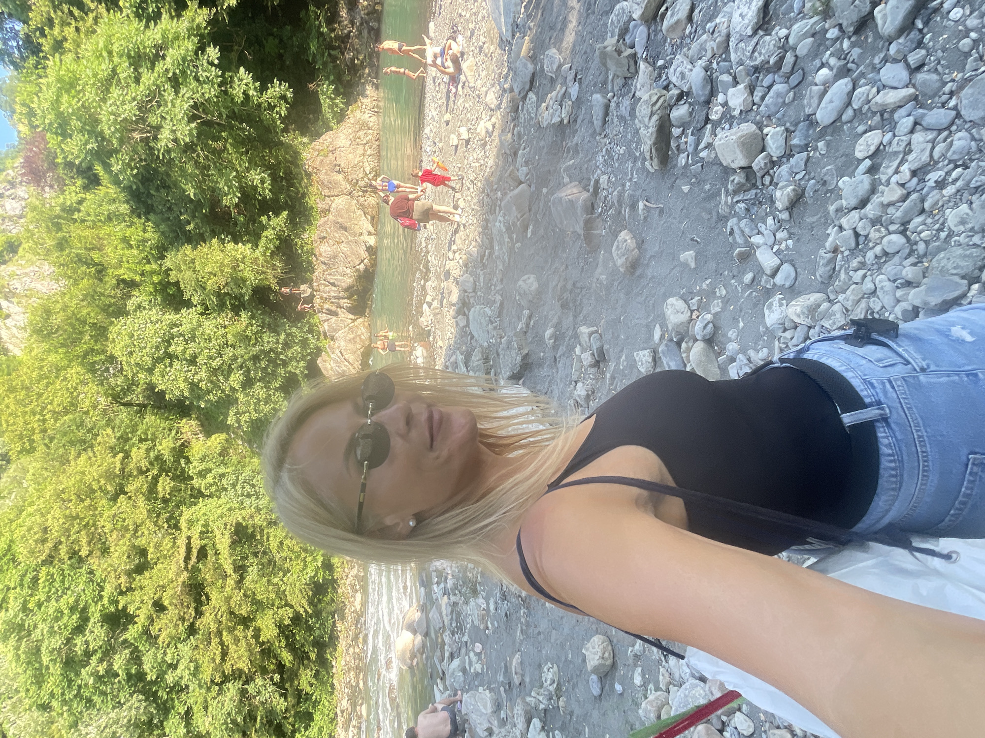 A woman with blonde hair and sunglasses poses for a selfie near a rocky riverbank. She is wearing a black tank top and blue jeans. In the background, a group of people are swimming and relaxing in a turquoise river. The river is surrounded by lush green trees and rocky cliffs. The scene appears to be a popular spot for outdoor recreation. Based on the vegetation and the presence of a river with rocky banks, this location is likely a natural area within or near Sochi, Russia, known for its scenic river gorges and outdoor activities.