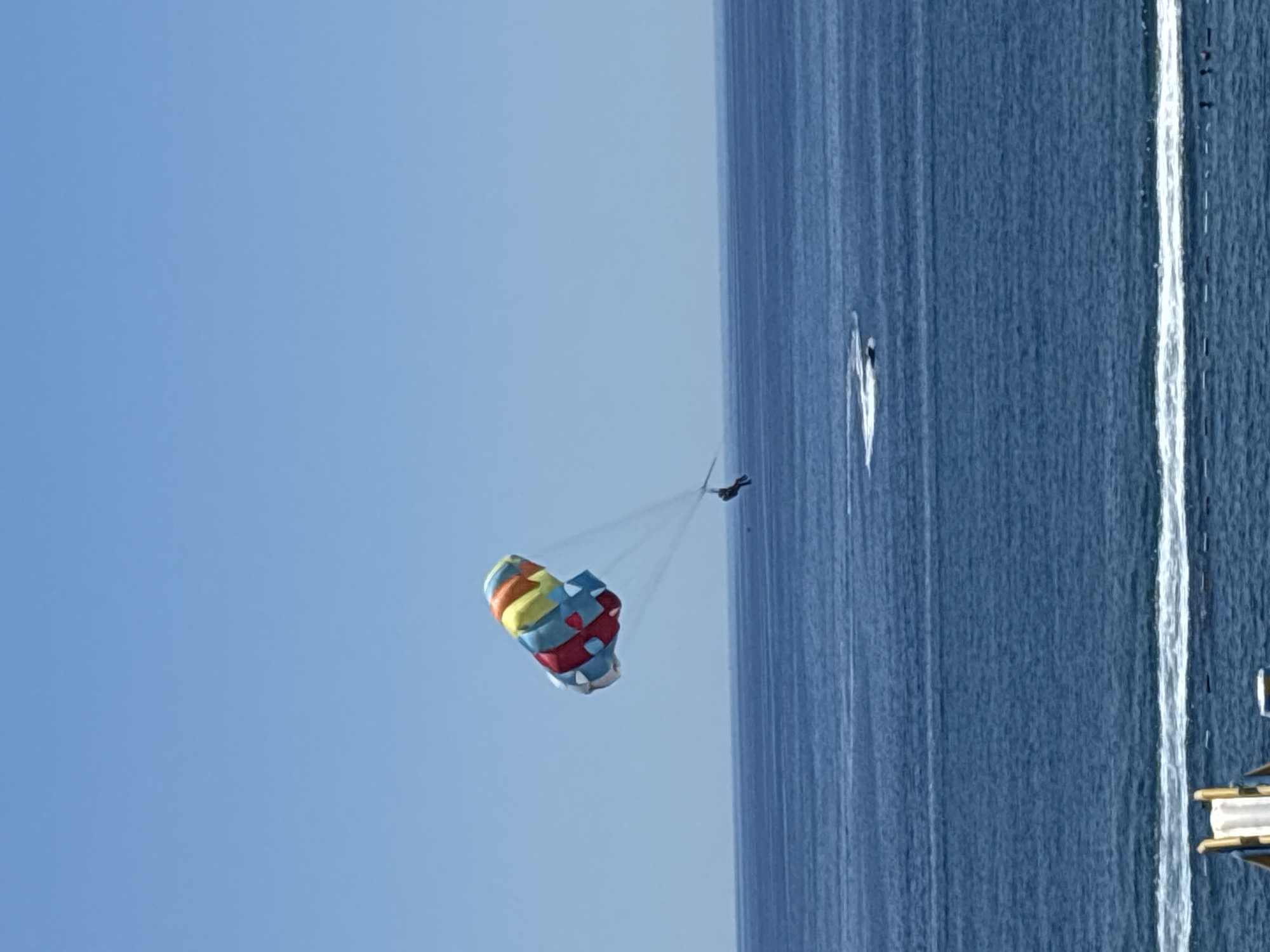 The image shows a parasailer being pulled by a boat over the Adriatic Sea. The parasail is colorful, with segments of red, yellow, blue, and white. The sea is a deep blue, with white wakes from the boat and possibly other watercraft visible. The sky is clear and blue. In the foreground, the edge of a beach or pier can be seen, with part of a white structure, possibly a slide, visible. The location is the coast of Boreti, Montenegro.