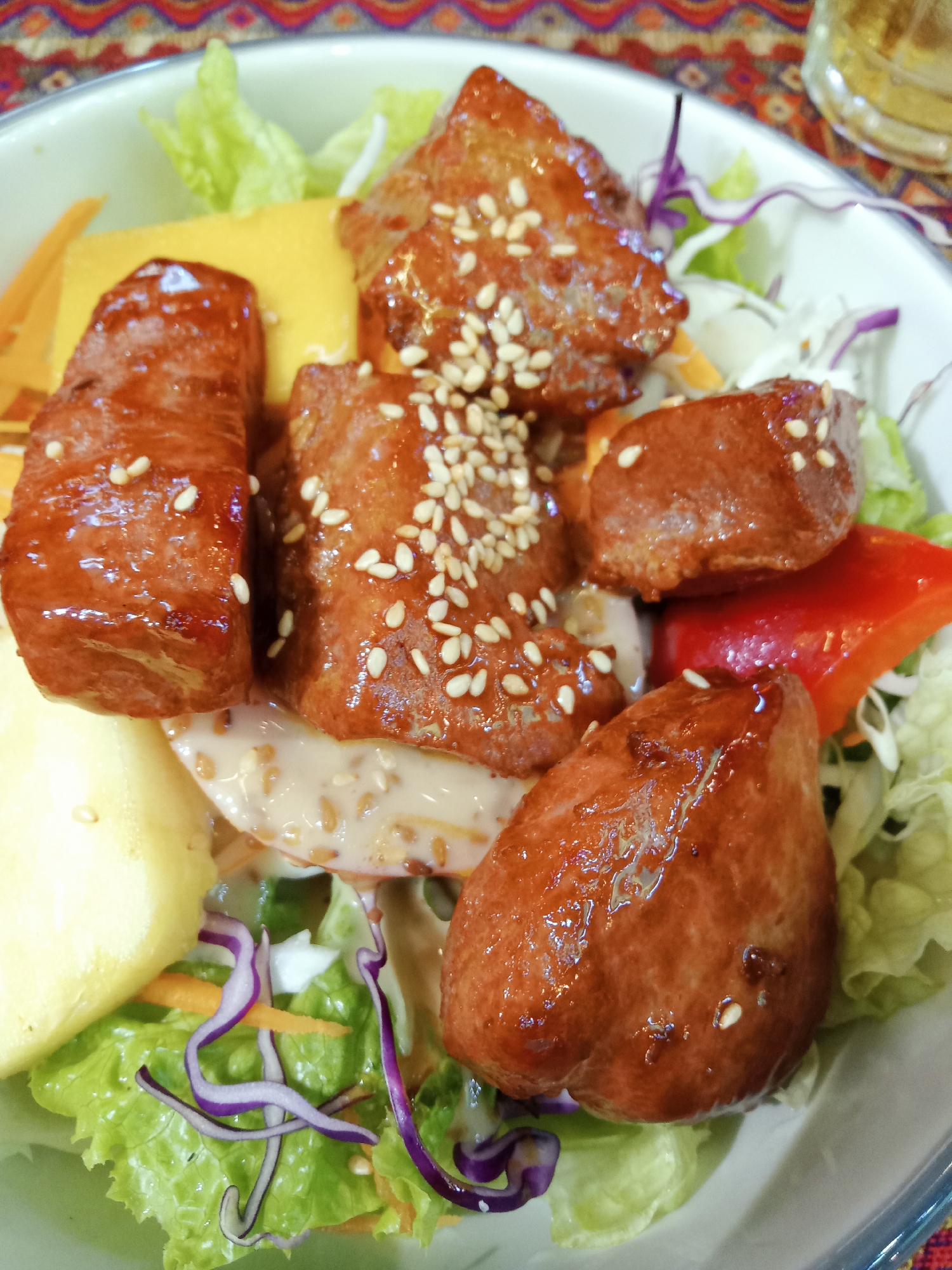 A close-up shot depicts a salad in a white bowl. The salad contains lettuce, shredded carrots, red cabbage, yellow peppers, and pieces of glazed meat, likely pork or chicken, sprinkled with sesame seeds. A creamy, speckled dressing is visible on the salad. The bowl sits on a patterned tablecloth. Based on the description provided, this photo could have been taken in Nha Trang Ward, Vietnam.