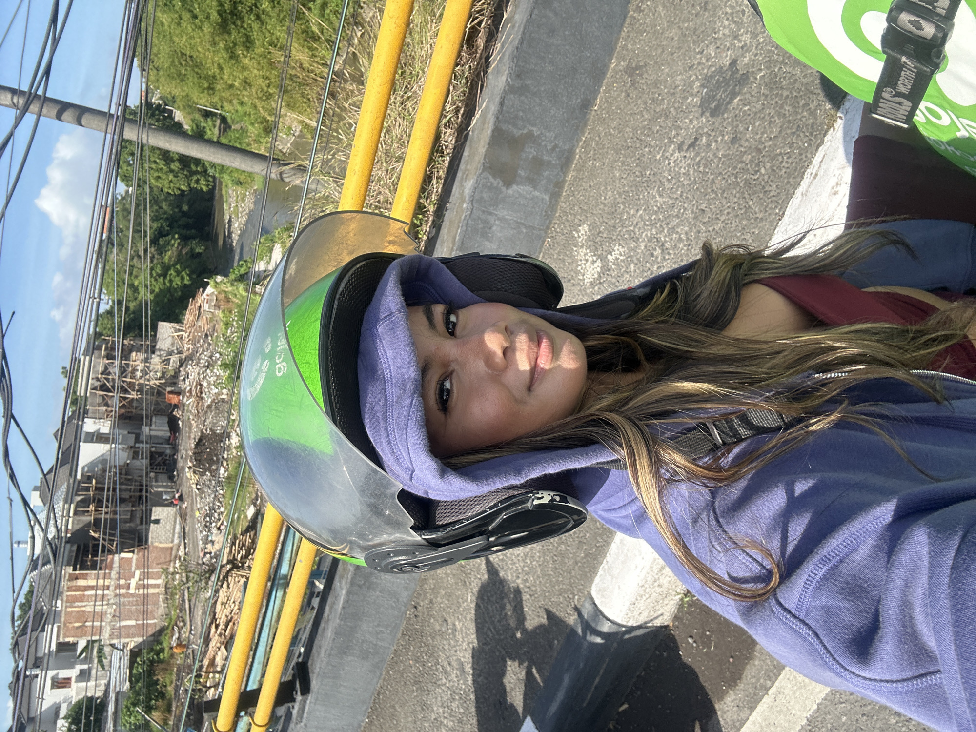 A woman with long, light brown hair and brown eyes is pictured in a selfie from the shoulders up. She is wearing a purple hooded sweatshirt and a green and clear motorcycle helmet. She is sitting on a motorcycle, as indicated by the helmet and the green bag with the Gojek logo visible to her right. The background shows a street with yellow railings and a river with trees on either side. In the distance, a building is under construction. The scene appears to be in Denpasar, Indonesia, as suggested by the Gojek logo, a ride-hailing service popular in the region. The sky is blue with some scattered clouds. The lighting indicates it is daytime, likely mid-morning or afternoon.