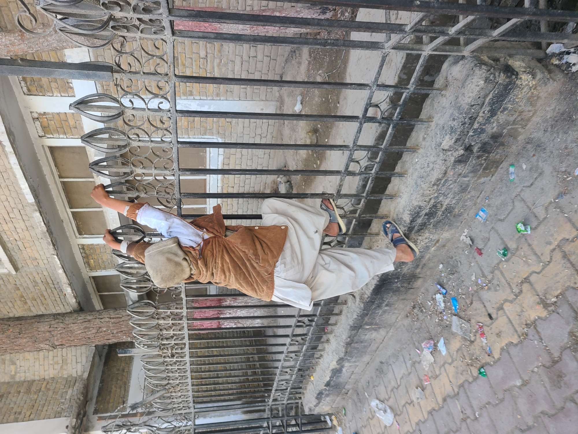 A person wearing a brown jacket, white shalwar kameez, and a grey hat is holding onto a black metal gate. The gate is in front of a building constructed of yellow bricks. The ground is paved with patterned bricks and is littered with debris. The image was likely taken in Quetta City Tehsil, Pakistan.