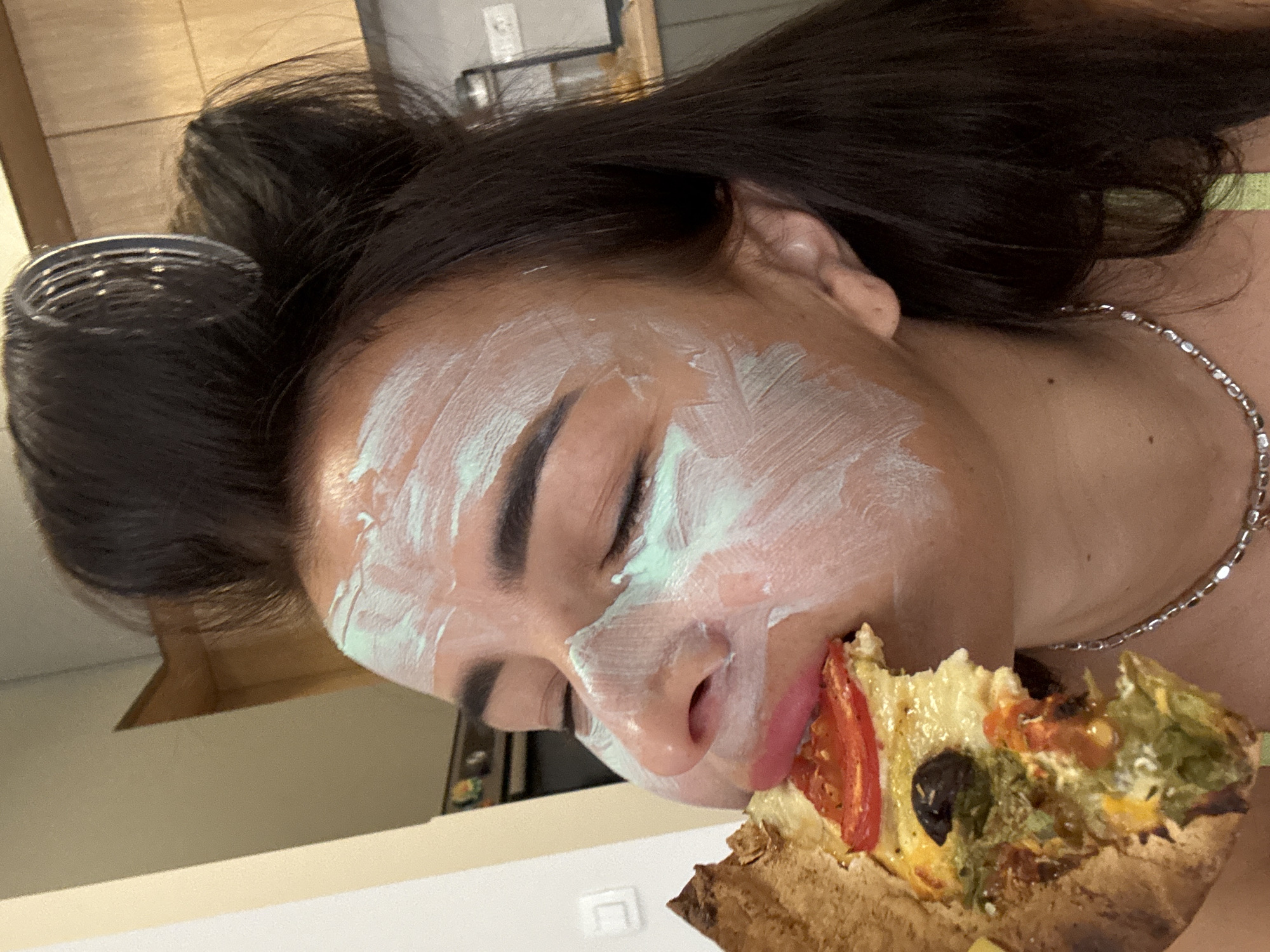 The media file shows a close-up, tilted-down view of a person with dark hair, wearing a white face mask with green undertones, and eating a slice of pizza. The person's eyes are closed, and their lips are painted pink. They are wearing a silver necklace. The pizza appears to have various toppings, including tomato slices and black olives. The background is blurred, but hints of a kitchen or dining area with wooden cabinets and some shelving are visible. The lighting suggests it could be daytime, but no specific time can be determined. There is no visible text in the image. The overall impression is one of casual self-care combined with a snack.