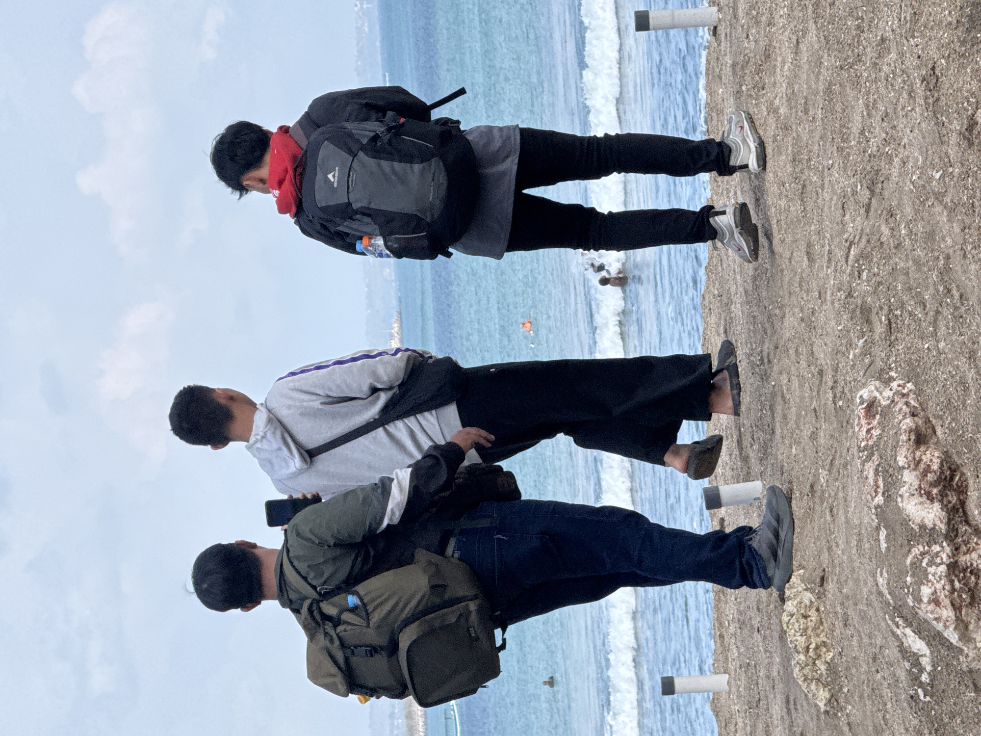 Three young men, appearing to be in their late teens or early twenties, stand on a sandy, rocky embankment overlooking a body of water, likely the ocean in Kuta, Indonesia, given the context of the prompt. The sky is overcast. The man on the left wears a dark green jacket, blue jeans, and dark grey sneakers, carrying a large, olive-green backpack. He is holding a black smartphone in his right hand. The middle man wears a grey hooded sweatshirt with purple accents on the sleeves, black pants, and black flip-flops. He has a dark bag slung across his body. The man on the right wears a black jacket over a grey t-shirt, black pants, and grey sneakers. He carries a large black backpack with a red hood peeking out from the top. Both the middle and right men are looking out towards the water. In the water, several figures are visible, some swimming and others wading. The embankment has several white posts with dark caps, typical of beach infrastructure. The scene appears to be a casual outing by the sea.