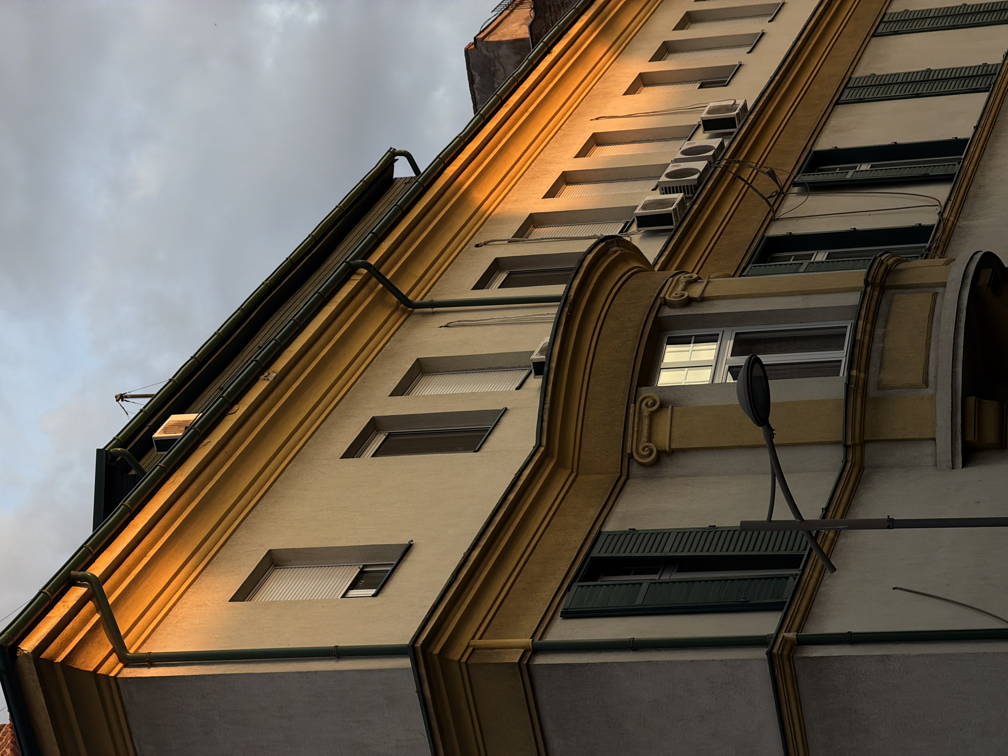 The image depicts an upward-angled view of a tall building in the Vracar Urban Municipality, Serbia. The building has a beige facade with golden architectural details. Windows of varying sizes are visible, some with open blinds. Air conditioning units are mounted on the facade between some windows. A decorative architectural curve is present near the center of the image. A streetlight extends from the building's facade. The sky is overcast.
