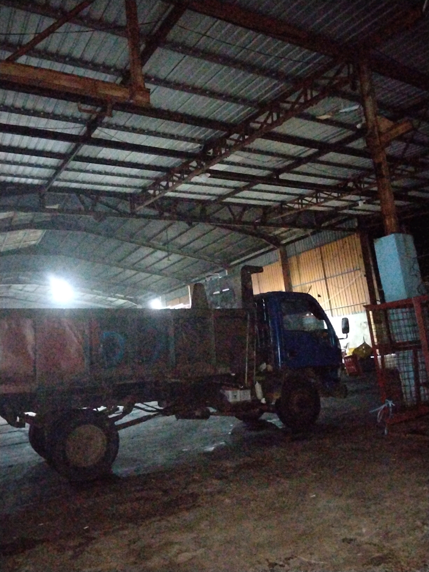 In the city of Moro, Indonesia, a parked blue truck with a damaged cargo bed sits inside a dimly lit warehouse or workshop. The corrugated metal roof of the structure is supported by a network of dark metal beams and wooden posts. Bright lights are visible in the background, suggesting an open doorway or windows. The truck's tires are visible, and its overall condition appears weathered and used.