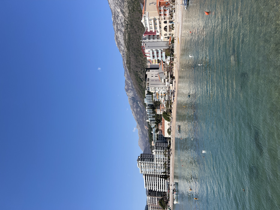 This image captures a panoramic view of the developed coastline of Budva, Montenegro, specifically a section of its beach and bay, backed by mountains under a clear sky.
The foreground consists of calm, clear, turquoise water, exhibiting gentle ripples. Numerous mooring buoys are visible: approximately six white buoys, one orange buoy in the lower right, and one yellow buoy in the lower-left mid-ground. Several small boats are moored in the bay; two white boats are positioned towards the left, another smaller dark boat is centrally located, and a fourth light-colored boat is partially visible on the far right.
Beyond the water lies a light-colored sandy and pebble beach, which appears largely devoid of individuals. A dense line of multi-story buildings forms the immediate backdrop to the beach. On the left, several modern, white high-rise buildings, approximately 7 to 10 stories tall, feature numerous balconies. Towards the center and right, the buildings vary in style and color, including structures with white, beige, red, and yellow facades, some displaying pitched roofs, arches, and balconies. These buildings appear to be a mix of residential, hotel, and commercial establishments, with some ground floors suggesting cafes or restaurants. Palm trees are sparsely visible among the buildings.
The background is dominated by a substantial mountain range. The lower slopes of these mountains are covered with sparse, possibly deciduous, vegetation or light scrub, while the upper reaches are rocky and appear more barren. The sky above is a uniform, deep blue, with a single small, wispy white cloud present in the upper-center right portion of the frame. The scene is illuminated by direct sunlight, indicating daytime conditions. No individuals are discernibly present on the beach, in the water, or on the boats.