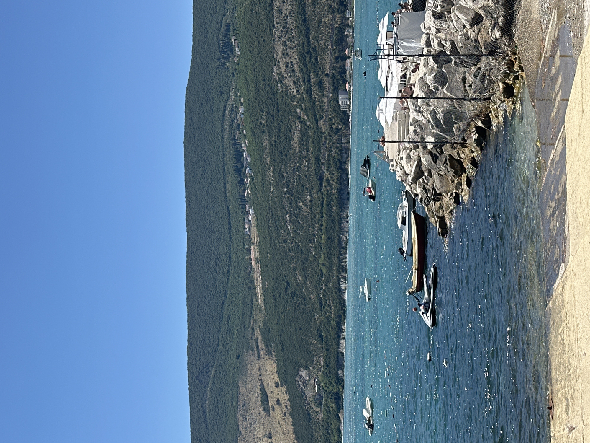 This is a daytime, eye-level shot of a coastal scene in Igalo, Montenegro, under a clear blue sky. The foreground features a concrete pathway leading into shallow, calm, turquoise water. To the right, a rocky jetty juts out into the sea, adorned with white lounge chairs, umbrellas, and a few people relaxing or walking. Further out in the water, several boats are moored or navigating: a jet ski is in motion, and other small vessels are visible, including a sailboat. In the background, a lush, green, tree-covered hill rises steeply, with a scattering of houses and buildings visible on its slopes. The water is slightly choppy, with sunlight glinting off its surface.