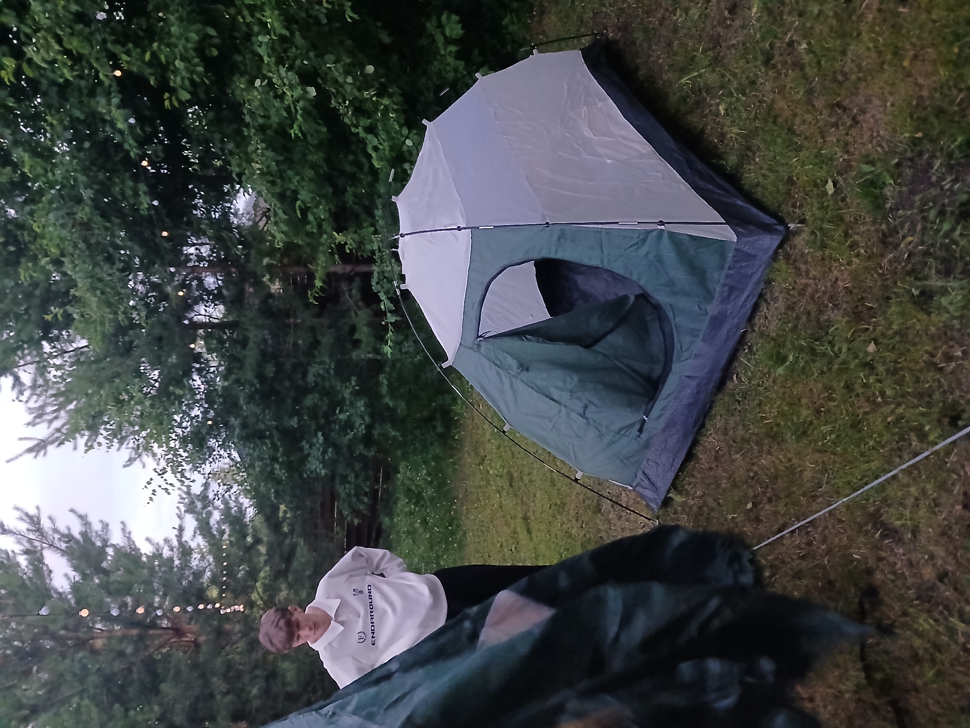 A young male, appearing to be in his late teens, stands outdoors in a grassy area surrounded by trees. He is wearing a white collared shirt with dark pants. To his right, a two-toned green and white tent is set up on the grass. In the background, strings of lights are visible, possibly strung between trees. The sky is overcast. The scene appears to be in Verkhneye Dubrovo, Russia.