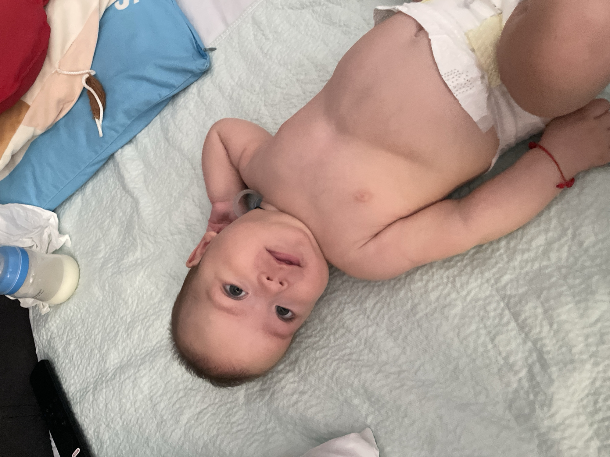 This media file shows a close-up, high-angle shot of a baby lying on a textured, light blue surface. The baby is positioned in the center of the frame, lying on its back with its head turned to the right. The baby is wearing a diaper and appears to be very young. The baby has light skin and brown hair, and its eyes are open and looking towards the camera. The baby's right arm is bent at the elbow, and its hand is near its head. A red string bracelet is visible on the baby's left wrist.
To the left of the baby, there is a partially visible baby bottle with a blue nipple, containing a white liquid. Further to the left, out of focus, are some colorful fabrics and a blue pillow. A dark object, resembling a remote control, is also visible at the bottom left of the image.
The lighting appears to be soft and diffused, suggesting an indoor setting. The overall mood is calm and peaceful. No specific time of day or weather conditions can be determined from the image. No visible text is present in the image. The location context provided is Fălești, Moldova.