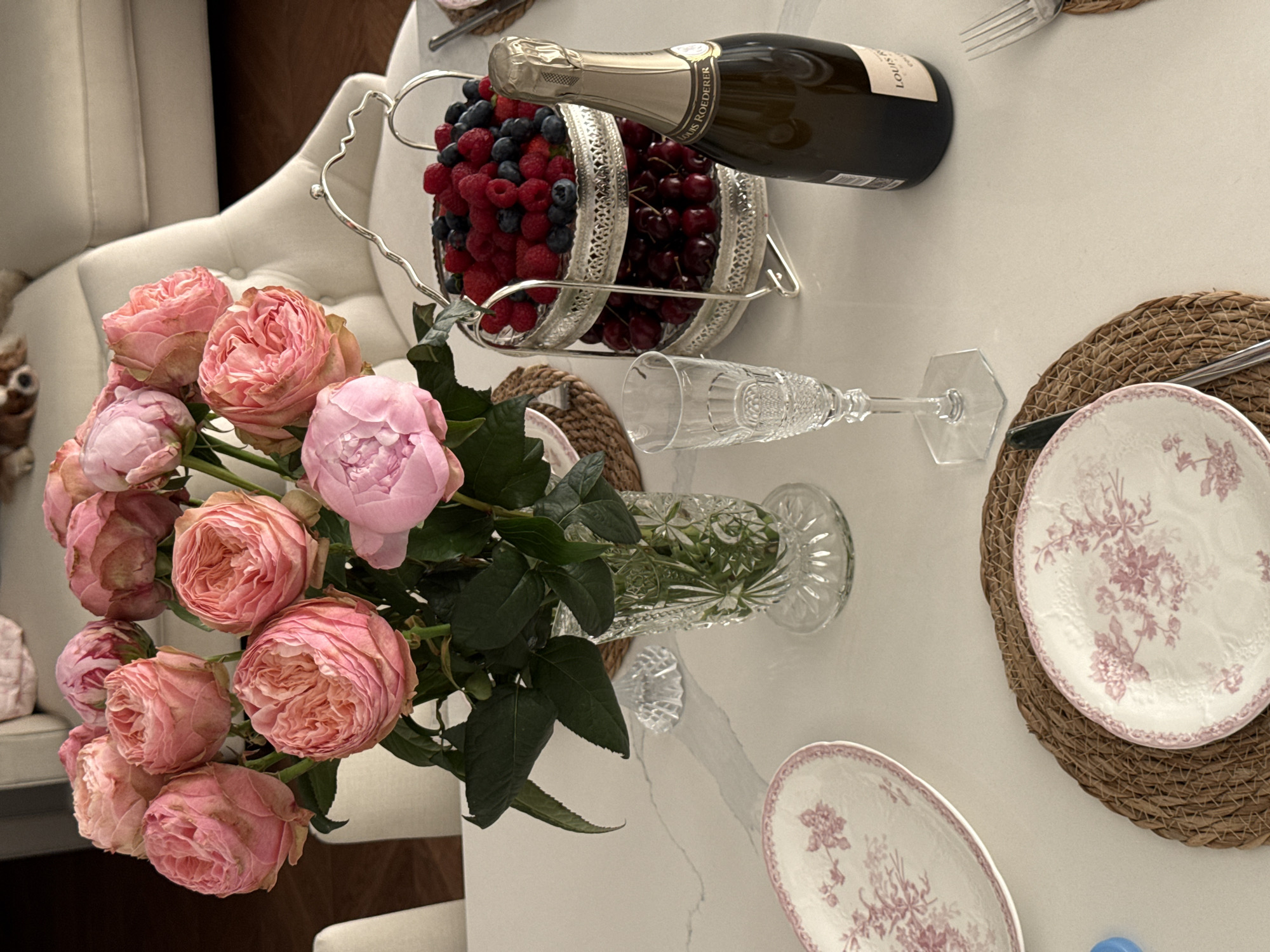 The image captures a detailed tabletop scene, likely set for a special occasion. It features a bouquet of pink roses arranged in a crystal vase, accompanied by a silver tiered stand filled with fresh berries, including raspberries, blueberries, and cherries. A bottle of champagne is horizontally placed next to the berries. A crystal champagne flute lies on the table. Two plates with a floral pattern are set on woven placemats. A fork and knife are visible near the plates. The setting suggests a refined and celebratory atmosphere. There's no evidence to suggest it's located in Moscow, Russia.