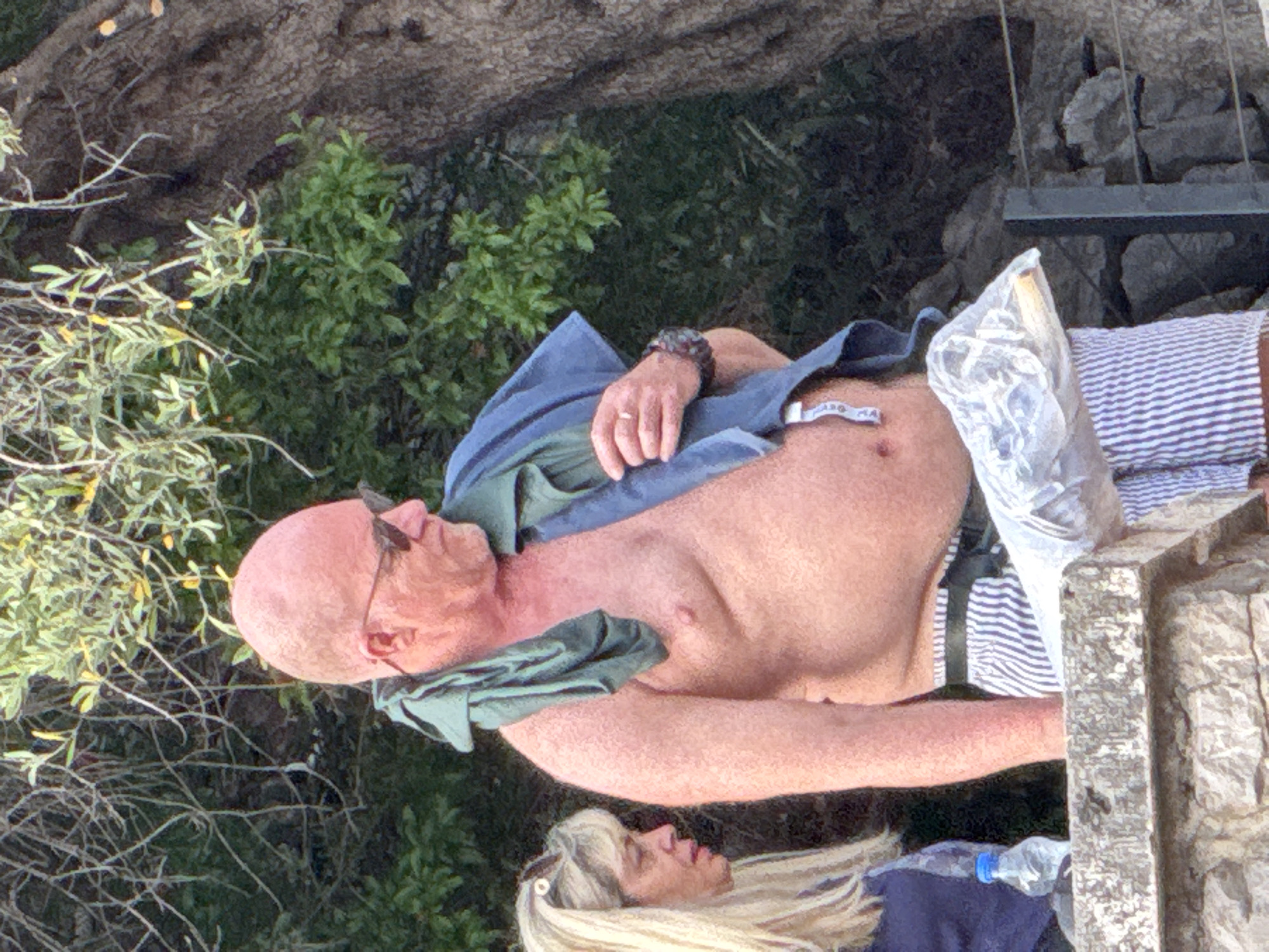 The image depicts a male and a female in Sveti Stefan, Montenegro, set against a backdrop of dense green foliage and a large tree trunk. The central figure is a fair-skinned, bald man with a prominent abdomen, wearing dark sunglasses, striped white and blue shorts, and a black belt. A green shirt is draped around his neck, and a blue towel is slung over his left shoulder. He wears a ring on his left ring finger and a watch on his left wrist, and holds a clear plastic bag containing an elongated light-colored object in his left hand. He is oriented towards the viewer's left. Partially visible on the left is a blonde-haired woman, wearing a dark top, looking downwards and to her right. A clear plastic water bottle with a blue cap is positioned in front of the woman. Both individuals are situated adjacent to a stone wall or railing in the foreground, with a dark metal railing partially visible behind the man on the right.