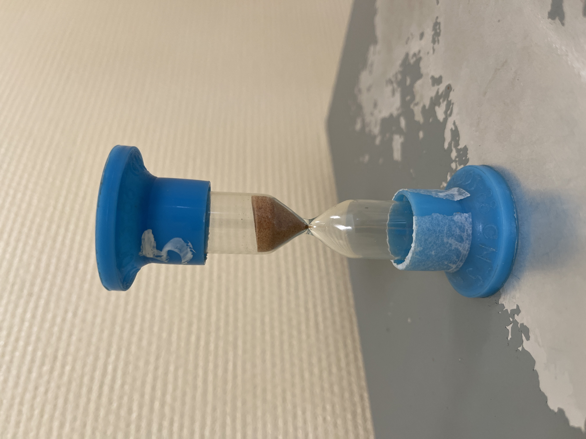 A blue plastic hourglass stands on a surface with peeling gray paint, next to a beige wall with vertical striped texture. The upper bulb of the hourglass is almost empty, while the lower bulb is nearly full with brown sand. Pieces of white tape are stuck on both ends of the hourglass. The location is not visible in the provided image to determine if it is in the city of Chaikovsky, Russia.