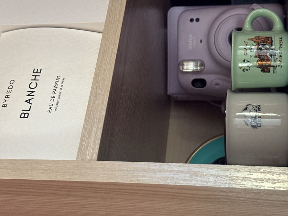 The image displays an indoor scene in the city of 3-Y Uchastok, Russia, featuring various objects arranged on and within wooden shelving. A light wood-grain shelf horizontally bisects the upper portion of the frame. Above this shelf, a white, curved-edge product package is visible, labeled with black text: "BYREDO", "BLANCHE", "EAU DE PARFUM", and "VAPORISATEUR NATURAL SPRAY". A vertical light wood-grain panel borders the left edge of the frame.
Below the shelf, positioned against a darker background, are three distinct items. On the far left, a partial view of a round, teal-colored object with a gold-colored rim is visible. Adjacent to it, a cream-colored mug with a dark brown rim displays a graphic of snow-capped mountains and the Cyrillic text "ОСЕТИЯ". To the right of the cream mug, a light green mug with a dark brown rim and handle features a multicolored illustration of an urban street scene, depicting buildings, a lamppost, and a white dog. This mug is inscribed with the Cyrillic word "ЗЕЛЕНОГРАВСК" at the bottom, and a partial word "CRAZ" accompanied by a heart symbol near the top. Behind these two mugs, a light purple Fujifilm Instax mini 11 instant camera is present, with its lens, reflective flash unit, and "instax mini 11" branding clearly visible. No individuals are present in the scene.