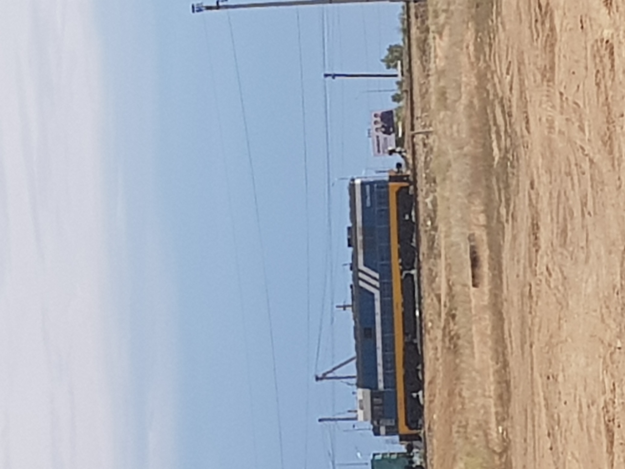 An outdoor, eye-level, long-shot photograph captures a blue and yellow train in Kurchatov, Kazakhstan. The train is positioned on railroad tracks and is the primary subject of the image. In the background, a billboard featuring three individuals is visible, along with a utility pole. The environment is dry and arid, with sparse, brown vegetation and dirt ground. The sky above is a light blue with some faint clouds. The overall impression is one of a vast, open landscape under a pale sky.