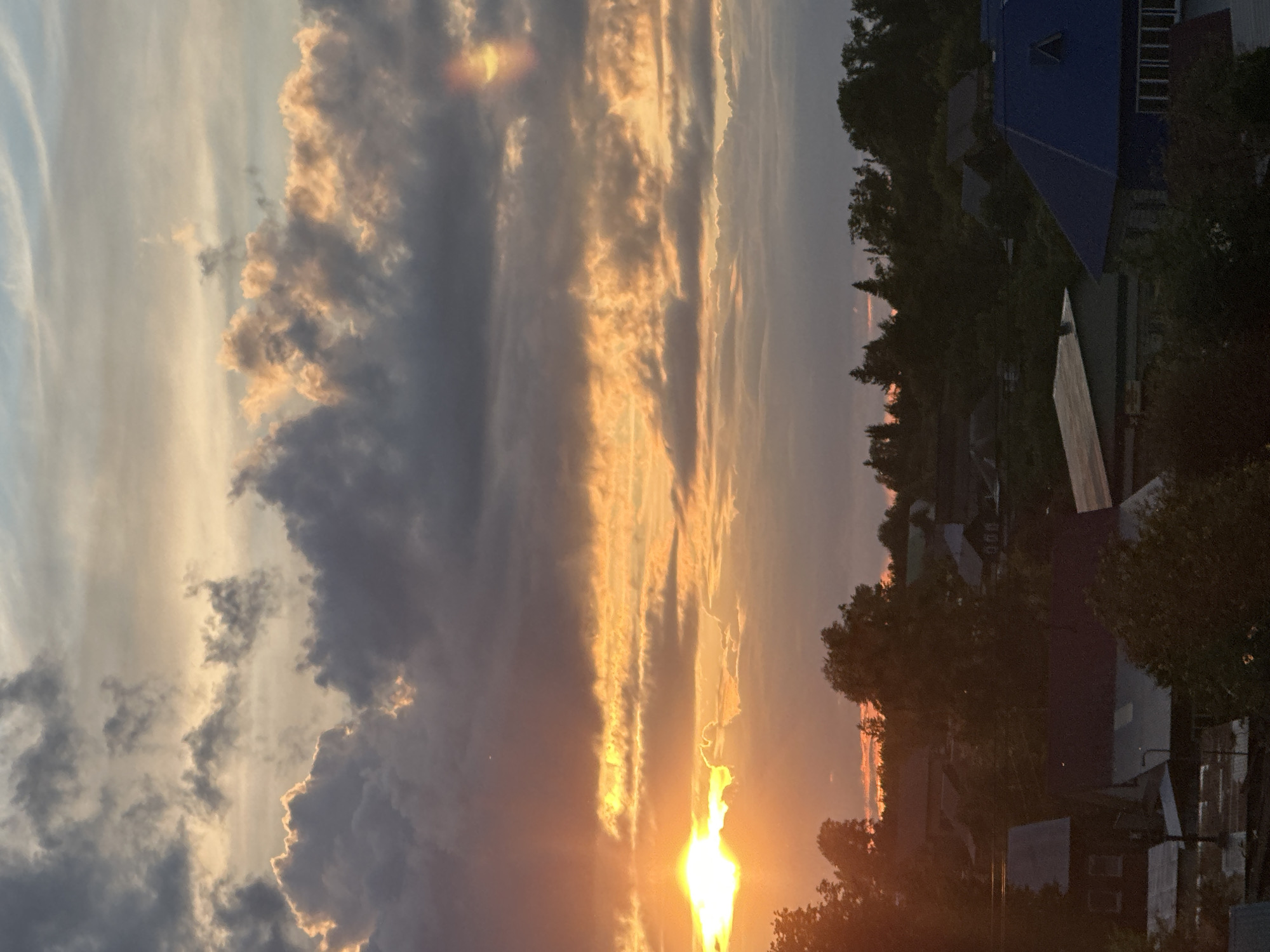 The image depicts a sunset in a residential area. In the foreground, the setting sun shines brightly between tree branches, illuminating the sky with orange and yellow hues. Above, clouds are streaked with these colors, transitioning to darker gray and blue tones. Houses and buildings are visible in the middle ground, with a blue roof standing out. The location appears to be a suburban or rural setting in Unknown, Unknown. No people are visible. The general scene is one of twilight over a residential area.