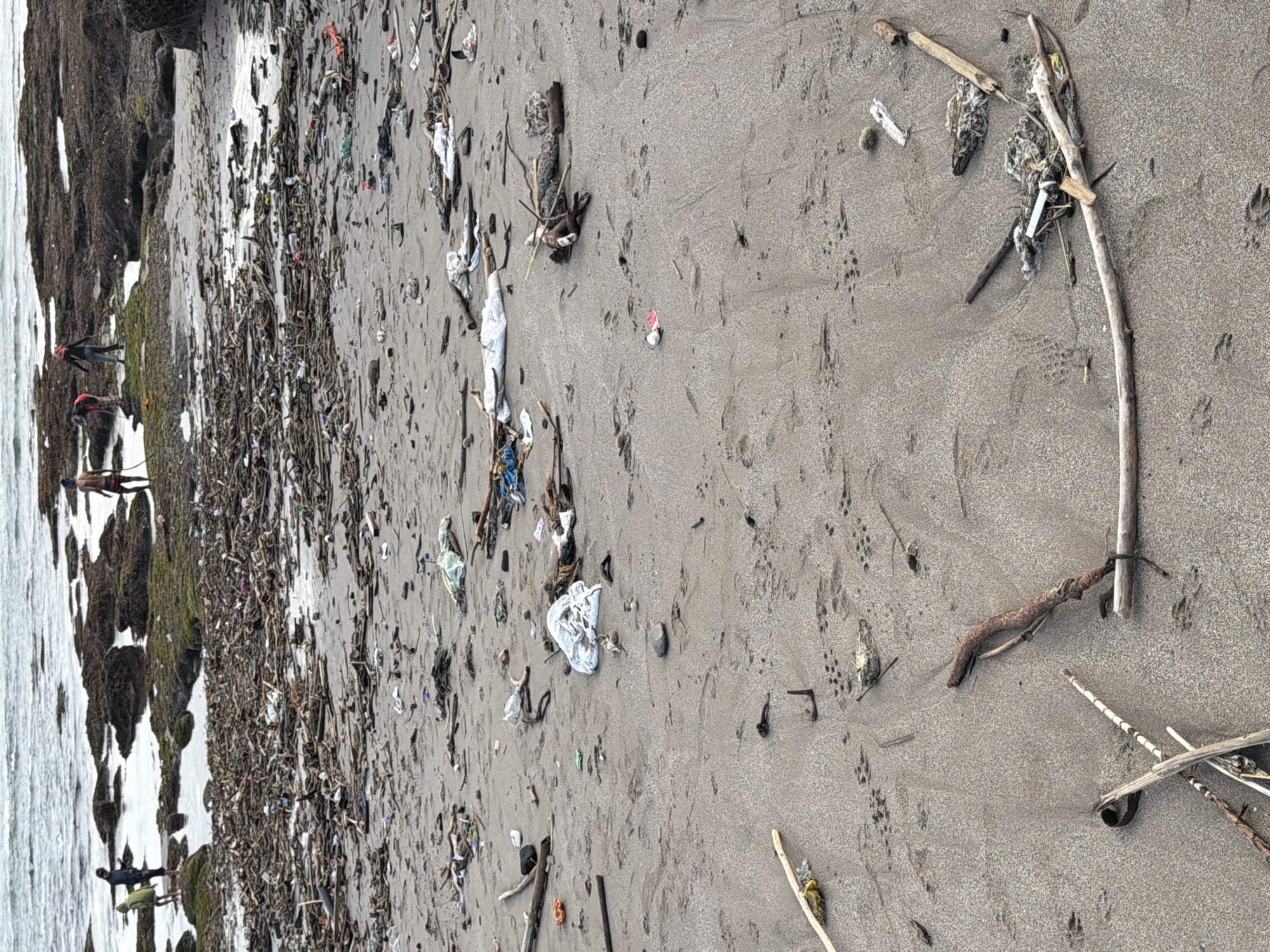 The image depicts a heavily littered beach in North Kuta, Indonesia. The foreground shows a sandy beach covered with debris, including driftwood, plastic bottles, bags, and other trash. Several sets of animal tracks are visible in the sand. In the middle ground, more debris is scattered across the beach and in the shallow water. In the background, a rocky shoreline is visible, with moss or algae growing on some of the rocks. The ocean water appears choppy. Three individuals are present on the rocky area in the background. One person, wearing a dark jacket and a green hood, stands on a rock. Another person, in a red top and dark pants, is bending down. A third individual, wearing a dark top and dark pants, is standing further to the right. The overall scene conveys a significant pollution problem on the beach.