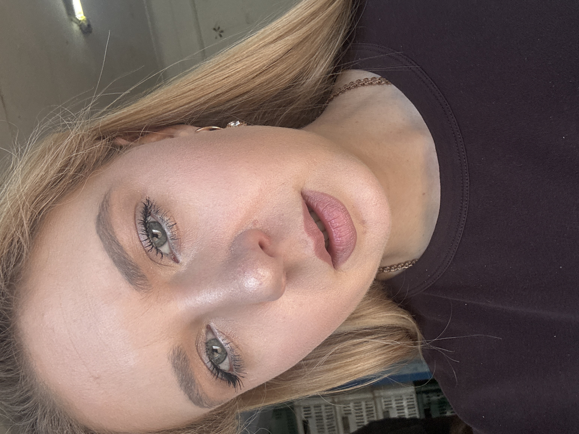A close-up, head-and-shoulders, selfie-style photograph shows a young Caucasian woman with blonde hair, fair skin, and green eyes. She is wearing a dark purple t-shirt and a gold chain necklace. A small, gold hoop earring with a clear gem is visible in her right ear. Her makeup includes mascara, eyeliner, eyeshadow, and a rosy-pink lipstick. The background is blurred and appears to be an outdoor setting with what looks like white crates or shelves. The lighting suggests it is daytime.