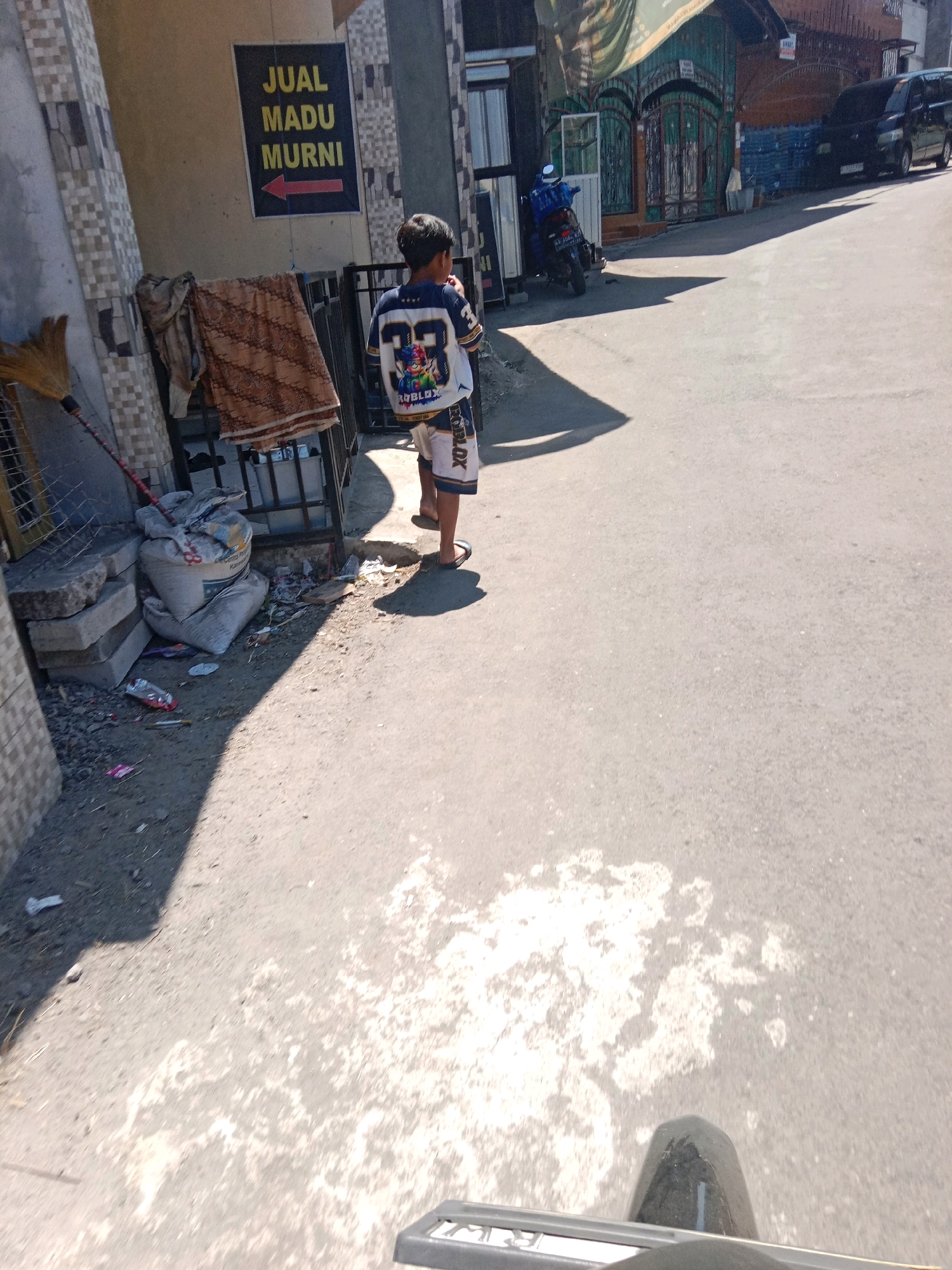 In Sumberdalem, Indonesia, on a sunny day, a young boy stands on a street. He is wearing a white t-shirt with blue accents and shorts with a Roblox graphic. He is facing away from the camera, looking towards a dark-colored motorcycle parked near a building. Next to the boy, a broom leans against a fence. A sign on the building reads "JUAL MADU MURNI" with an arrow pointing left, indicating a sale of pure honey. The street is paved and appears to be a residential area, with buildings and a dark-colored van visible in the background. The ground is littered with some debris and a large bag of what appears to be cement. The overall impression is a snapshot of everyday life on a street in a Indonesian town.
