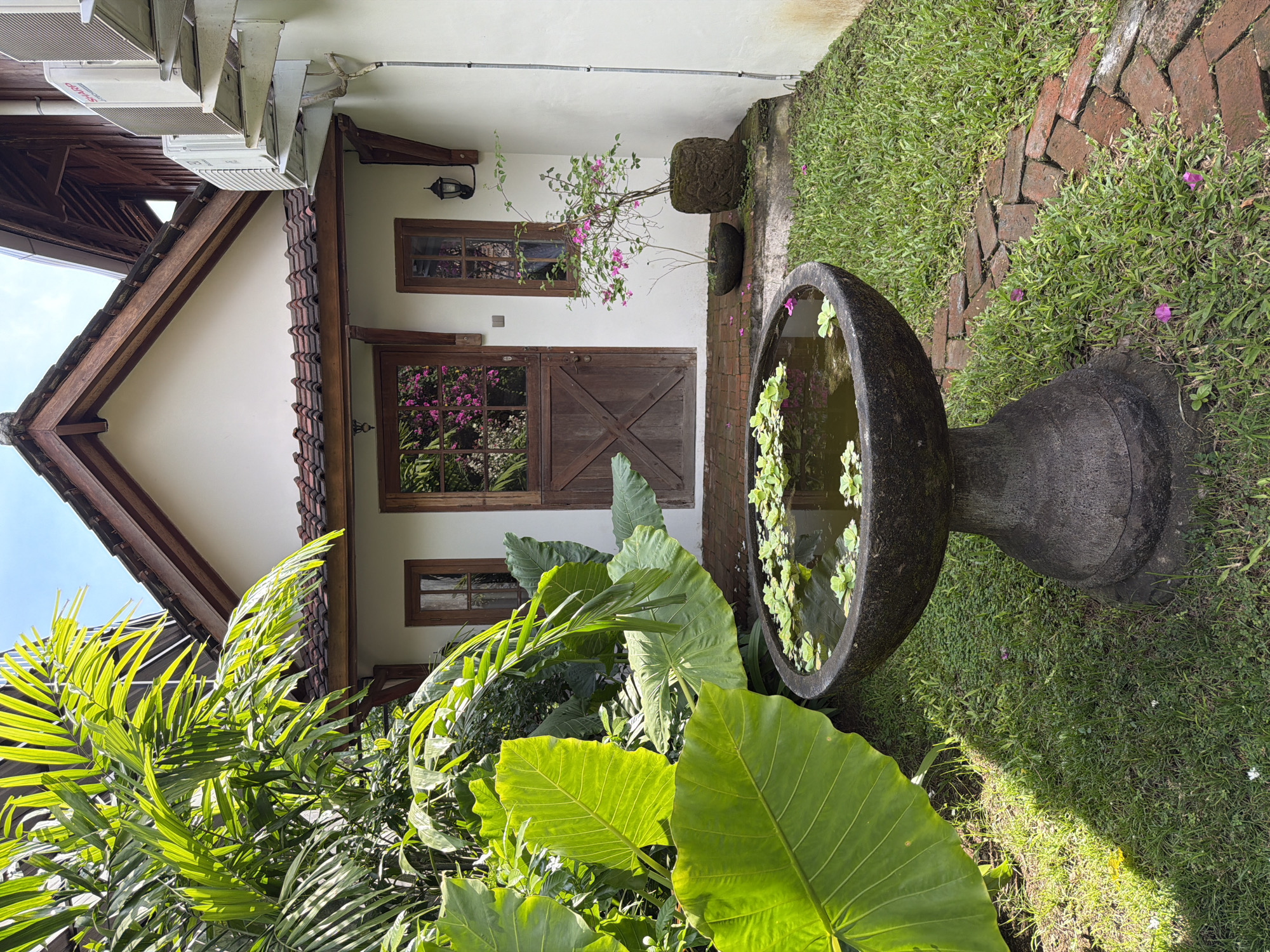 The image shows a building exterior in Kediri, Indonesia. The building is white with a brown roof and wooden trim. There is a wooden door with two windows above it. A light fixture is mounted near the top window. To the right of the door is a potted plant with pink flowers. A tall, stone birdbath filled with water and floating plants sits on the right side of the image. A brick path can be seen next to the birdbath. In the foreground, there are large green tropical plants. There is an air conditioning unit on the left side of the building. The sky is visible above the building.