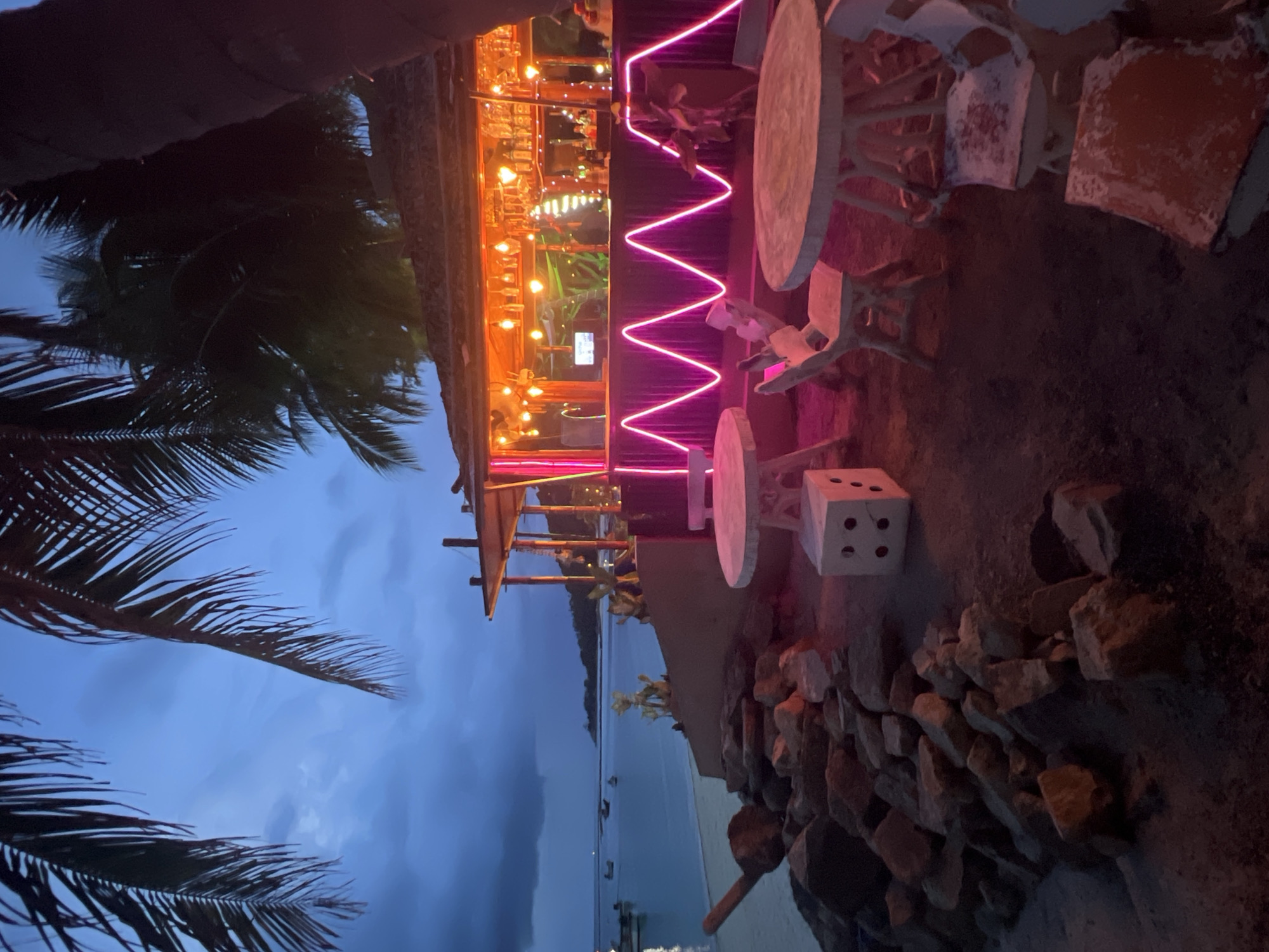 The image captures a dimly lit outdoor bar area on a beach in Ko Pha-ngan District, Thailand, during dusk. A palm tree overhangs the scene from the left, its fronds silhouetted against a twilight sky. In the background, the calm ocean stretches to the horizon, dotted with several small boats.
The bar itself is a rustic, open-air structure with a thatched roof, illuminated by numerous warm-toned lights. A prominent zigzag line of bright pink neon tubing decorates the front of the bar counter. Inside the bar, bottles of alcohol and glassware are visible on shelves.
In the foreground, several outdoor tables and chairs are arranged on the sandy, rocky ground. One table is round and light-colored, with three matching chairs around it. Another table, shaped like a cube with four circular indentations on its side, sits near a rocky embankment. The chairs appear weathered and are a light color, some with orange-tinted seats.
The environment is a tropical beach setting, with rough stones and sand forming the ground. The lighting suggests a transition from day to night, with artificial lights from the bar creating a vibrant contrast to the natural twilight. No people are clearly visible.