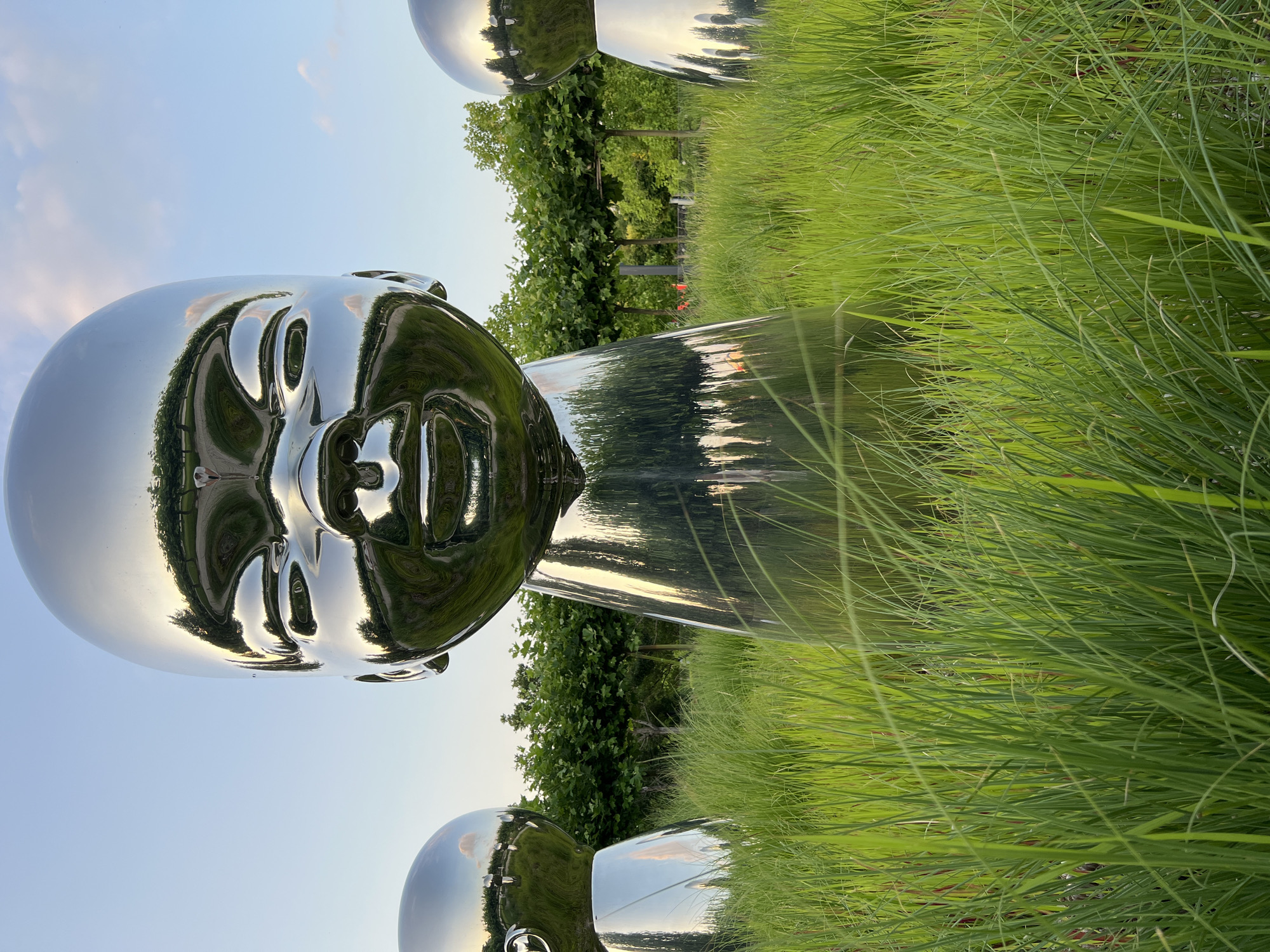 This media file features a large, chrome-plated sculpture of a human head, its reflective surface mirroring the surrounding environment. The sculpture is positioned outdoors, amidst lush green grass and trees, suggesting a park or garden setting. The sky above is a clear blue with some wispy clouds, indicating it is daytime, likely late afternoon given the warm light reflecting on the sculpture.
The sculpture's face is detailed, with discernible features like eyes, nose, and mouth, all rendered in a smooth, polished metal. The reflection shows trees, the sky, and possibly other sculptures or elements of the landscape. The orientation of the image is unusual, as it is rotated 90 degrees clockwise, making the sculpture appear to be standing upright rather than lying down.
The overall mood is serene and contemplative, enhanced by the natural setting and the abstract yet recognizable form of the sculpture. There is no visible text in the image. The location context provided is Krasnodar, Russia, which may imply the sculpture is part of an art installation or public display in that city. The scene appears to be a tranquil moment captured in an outdoor art space.
