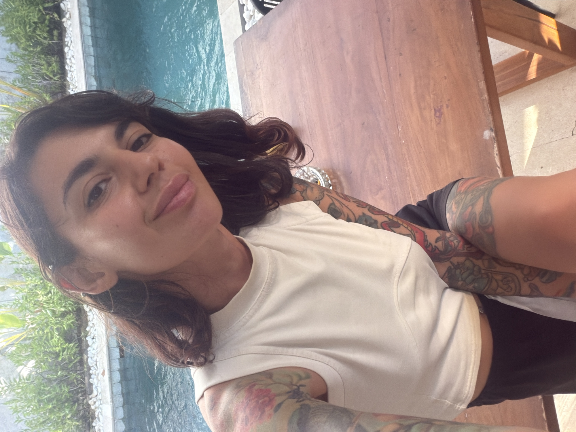 A medium-shot, slightly elevated selfie captures one individual seated outdoors next to a swimming pool in Kuta Selatan, Indonesia. The person, appearing female, has dark brown hair with a fringe, a light skin tone, and is smiling faintly. She wears a cream-colored sleeveless crop top and dark shorts. Both arms are extensively tattooed with multicolored designs, including floral motifs and abstract patterns. Multiple piercings, including an industrial bar and rings, are visible on her left ear. She is positioned at a rustic wooden table with visible wear and tear. To her left, an ashtray containing cigarette butts rests on the table. In the background, a clear blue swimming pool occupies the midground, bordered by white pebbles and lush green foliage under bright daylight conditions.