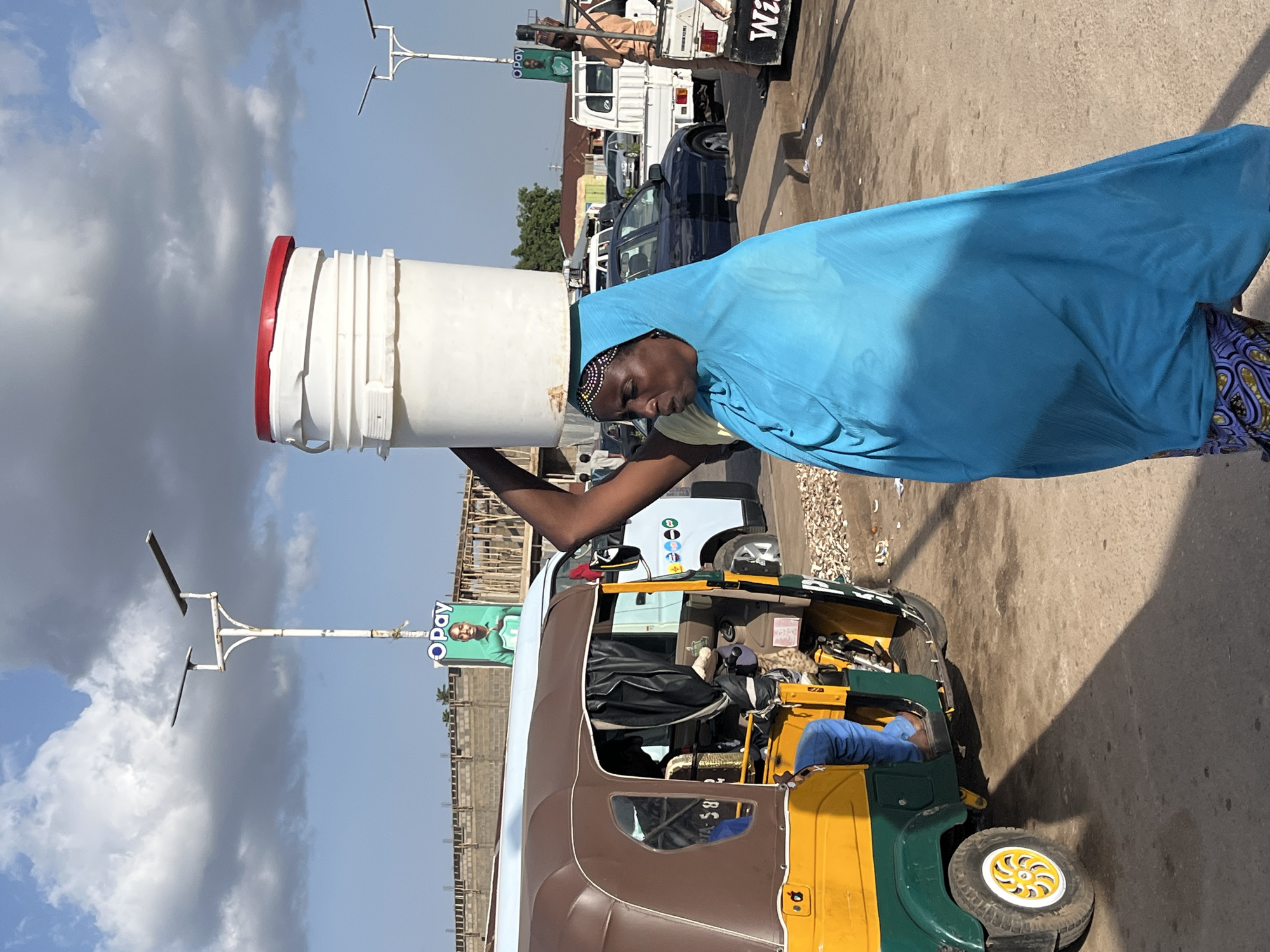 In Gura Top, Nigeria, a woman walks with a large white bucket, its red lid slightly askew, balanced on her head. She is draped in a vibrant blue cloth that covers her head and shoulders, with a patterned skirt visible beneath. To her left, a yellow and green auto-rickshaw is parked, its driver's legs clad in blue pants. The background reveals a street scene with a few cars, including a white truck with the word "Wi" partially visible on its side, and overhead, streetlights and billboards, one displaying "OPay" with a man's image. The sky is a mix of blue and dramatic clouds.