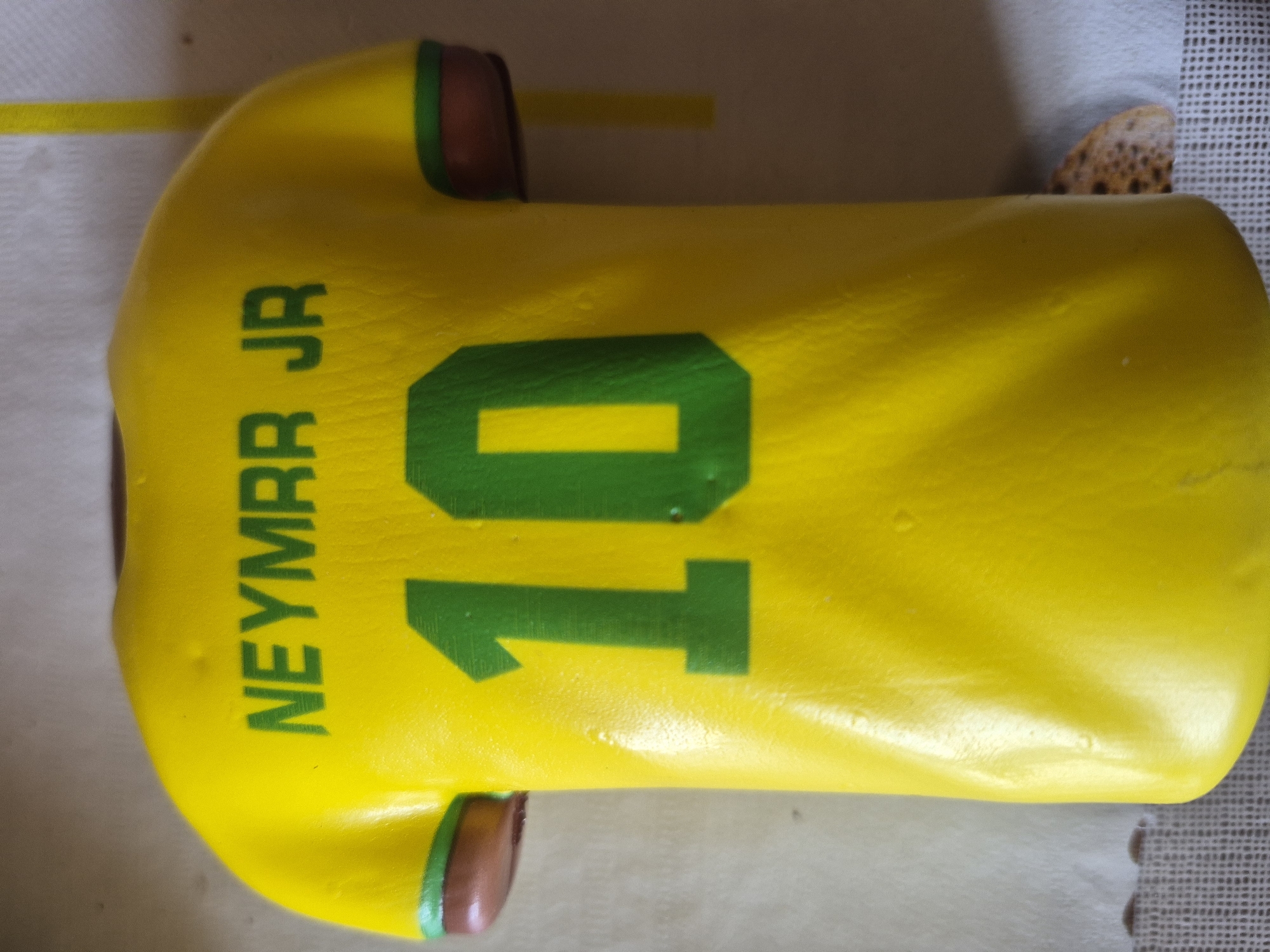 The image displays a cake intricately shaped and decorated to resemble a football (soccer) jersey, lying flat on a surface. The jersey is predominantly bright yellow, a color strongly associated with the Brazilian national football team.
Prominently featured on the back of the jersey is the name "NEYMARR JR" written vertically in green capital letters on the left side. To the right of the name, a large number "10" is displayed in matching green, signifying the jersey number. These details identify the cake as representing the kit of the renowned Brazilian footballer Neymar Jr.
The cake design includes short sleeves with brown and green trim, mimicking the cuffs of a real jersey. The surface of the cake appears smooth and slightly glossy, suggesting a fondant or similar edible coating.
The cake rests on a light-colored, possibly white or off-white, textured surface, which could be a tablecloth, baking mat, or serving tray. A thin, yellow stripe or ribbon is visible along the left edge of this surface. In the upper right corner, a small, irregularly shaped object with a speckled, brownish texture is partially visible. The overall scene suggests an indoor setting, likely a preparation or display area for the cake.
