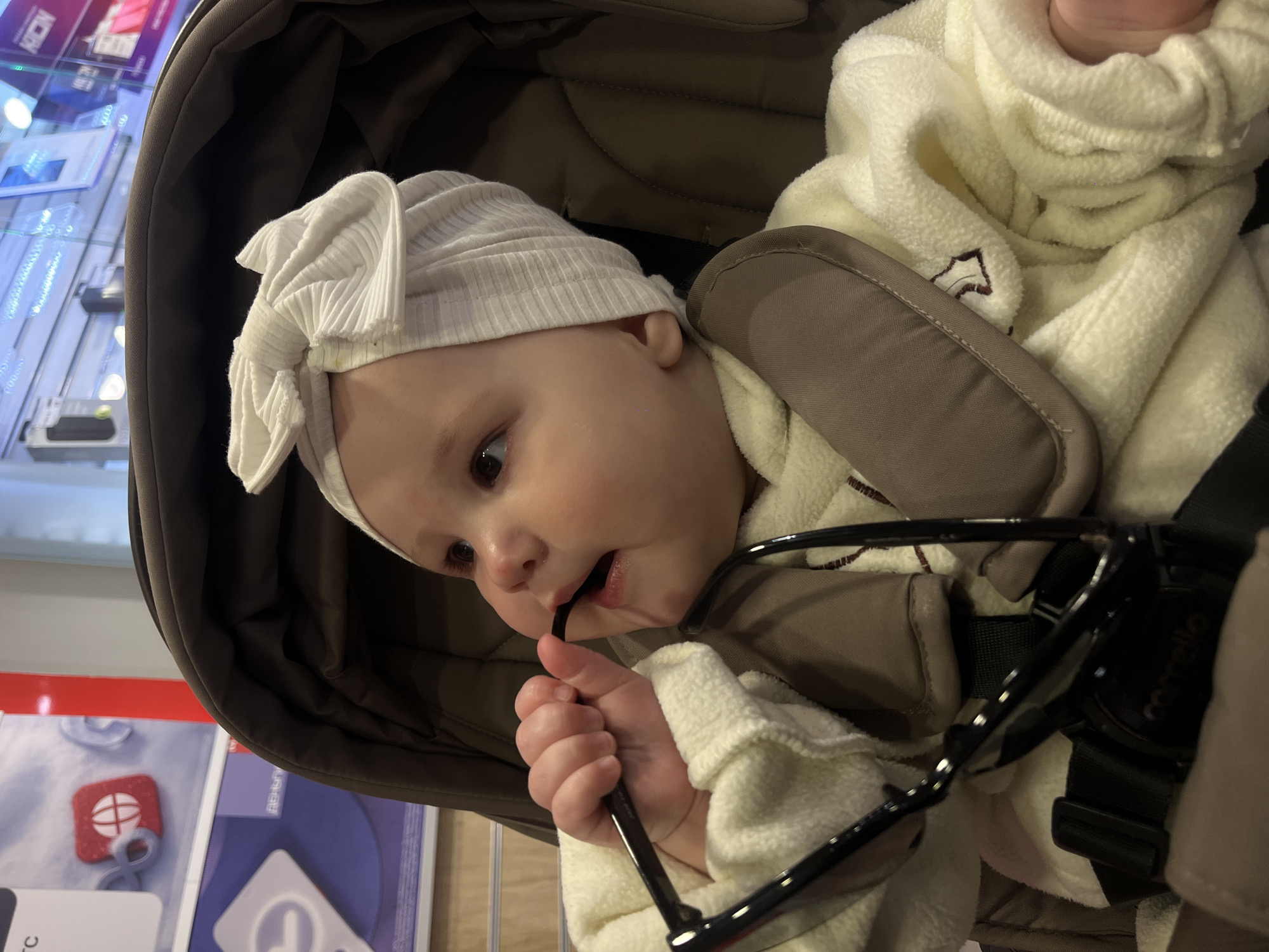 The image captures a baby, likely an infant or young toddler, situated in a brown and khaki stroller indoors, in what appears to be a retail store in Novopokrovskaya, Russia. The baby wears a light-colored, ribbed beanie with a decorative bow and a cream-colored, fleecy outfit. With a curious expression, the baby is actively engaged in holding and mouthing the arm of a pair of black-framed eyeglasses. The background, slightly out of focus and tilted, reveals illuminated display shelves stocked with various packaged products, some with visible text such as "KRON" and potentially "REDRAGON." The brand name "comello" is discernible on a padded strap of the stroller. The scene depicts a candid moment of a baby exploring their surroundings through play.