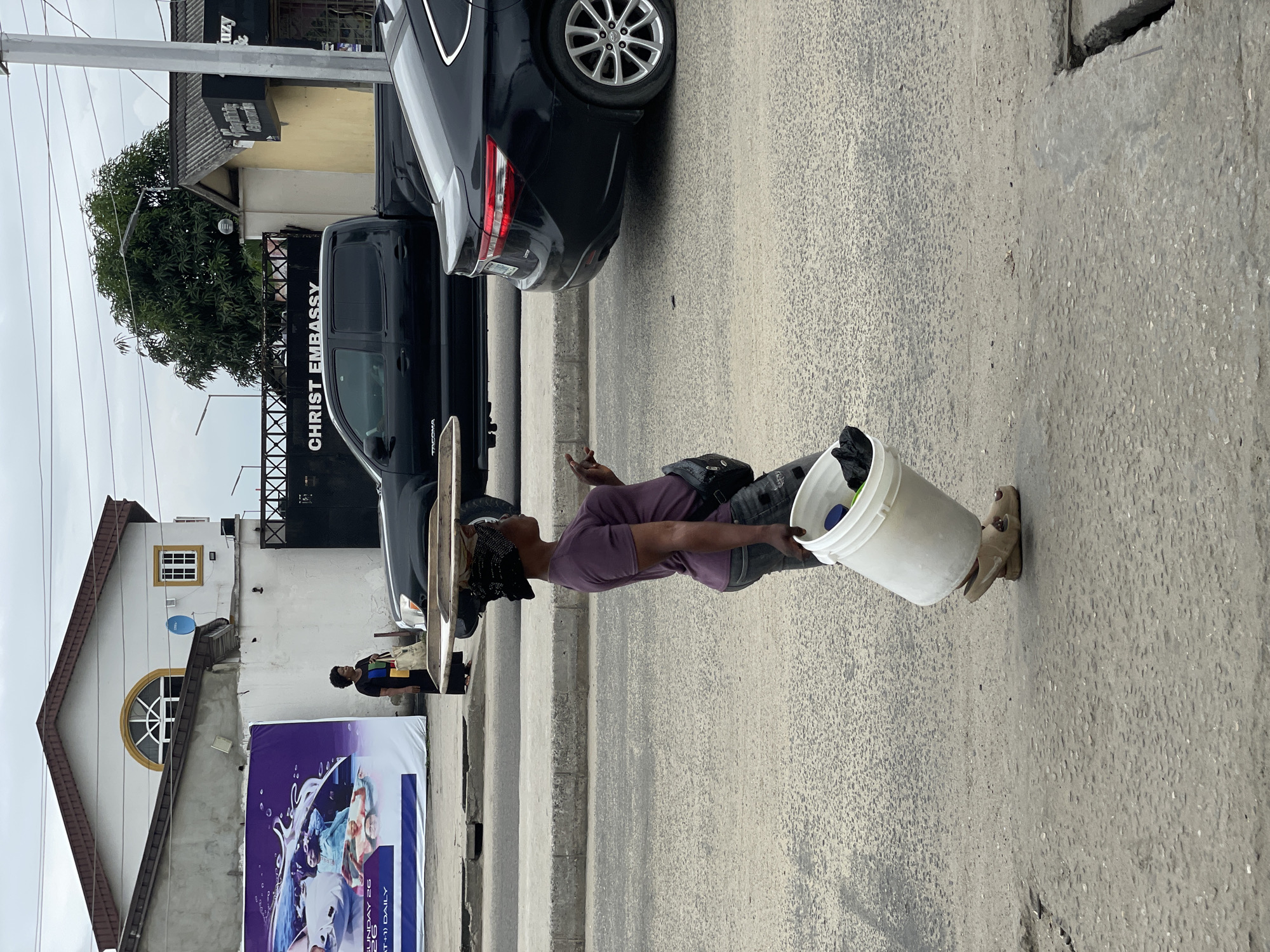 A woman walks down a street in Ado, Nigeria, carrying a large white bucket filled with black items in her right hand. She is also balancing a wooden tray laden with objects on her head, and wears a purple t-shirt, grey jeans, and beige sandals. A black crossbody bag is slung across her body. The street is paved with asphalt and concrete, and a grey concrete curb runs alongside it. In the background, a black pickup truck and a black sedan are parked. A white building with a brown roof, featuring a large arched window, is visible on the left side of the image, along with a large purple banner. On the right, a building with a yellow facade and a grey, corrugated roof has a sign reading "CHRIST EMBASSY" above a black gate. Overcast skies suggest a cloudy day.