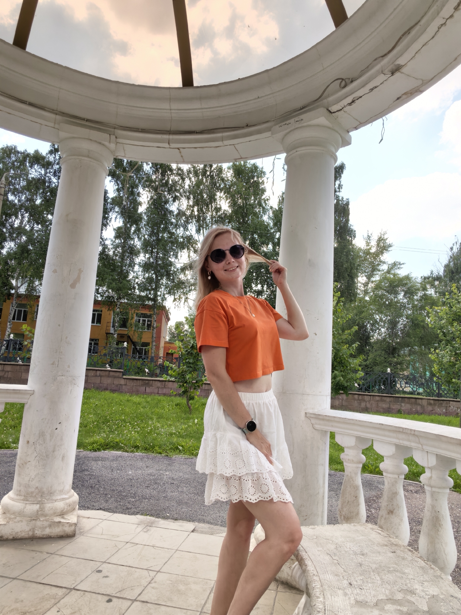 A young woman with blonde hair and sunglasses poses in an outdoor gazebo. She is wearing an orange crop top and a white skirt. She is holding her hair and smiling at the camera. The gazebo has white columns and a white railing. In the background, there are trees, a building, and a grassy area. The image appears to have been taken in Sterlitamak, Russia.