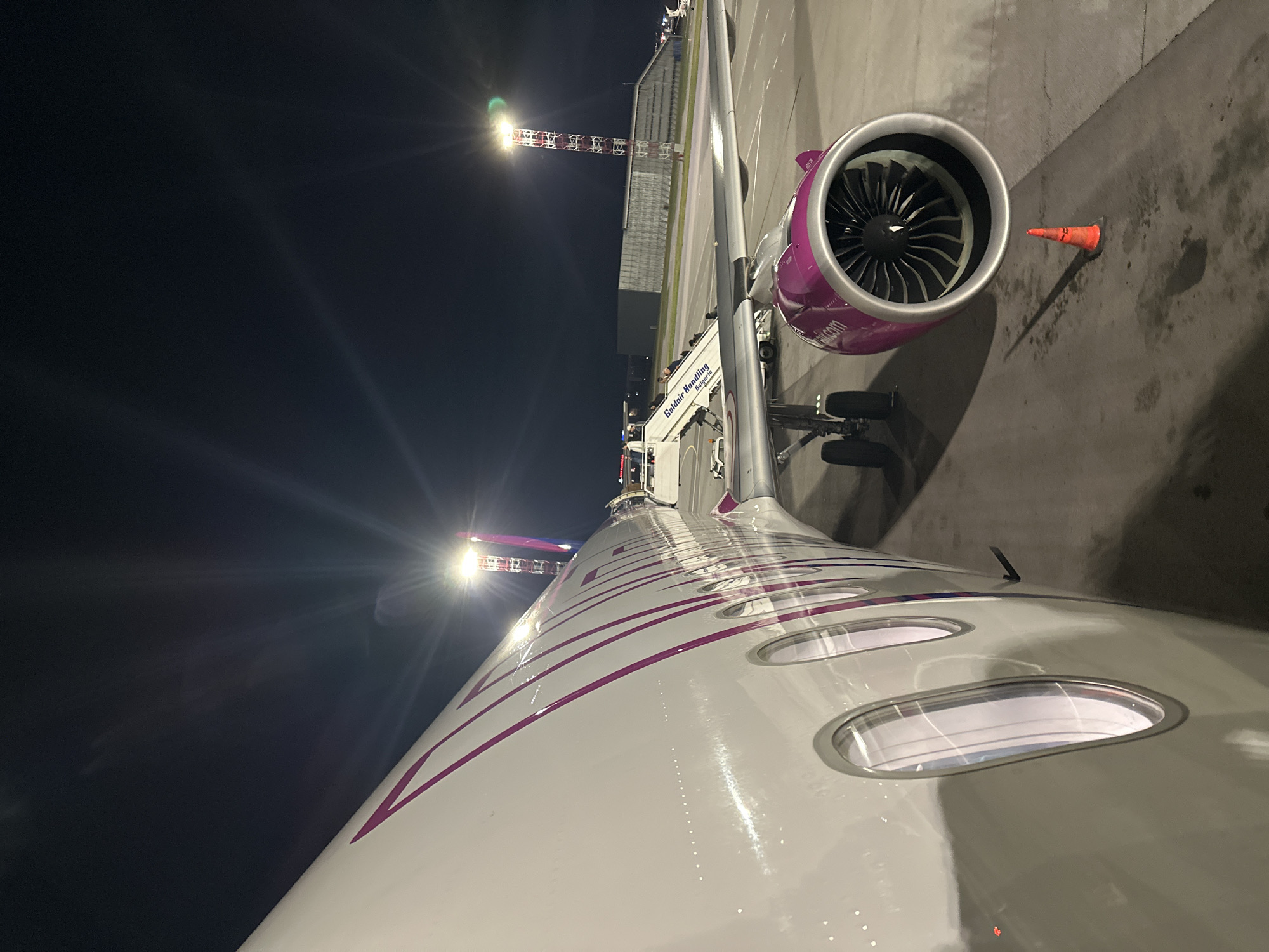 The image shows an airplane parked on an airport tarmac at night. The plane, painted white with purple accents, is connected to a passenger boarding bridge marked "Goldair Handling Bulgaria". A jet engine with a purple exterior is visible. An orange traffic cone is placed on the ground nearby. In the background, a floodlight tower is present. The location is likely Sofia Airport in Sofia, Bulgaria, based on the company logo on the bridge. No people are visible, though passengers may be inside the boarding bridge.