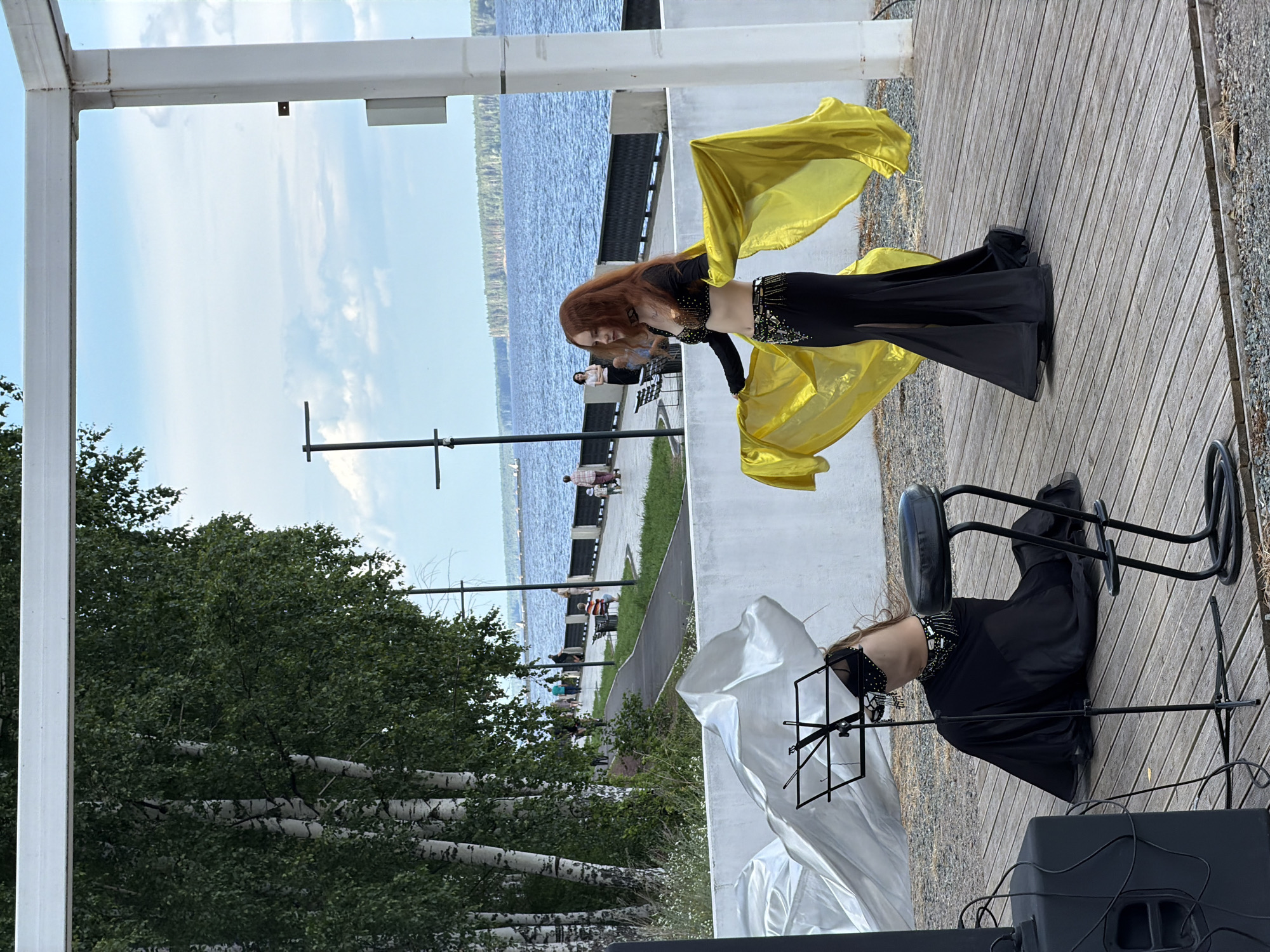 A woman with long, reddish-brown hair is performing a belly dance outdoors, on a wooden deck. She is wearing a black costume with a bejeweled top and a long, flowing black skirt. She is holding a large, flowing yellow veil, which she is moving expressively as part of her dance. The woman is angled away from the camera, with her head bowed slightly. To her right, a black chair with a metal frame is visible, and to her left, a music stand.
In the background, a body of water, likely a river or lake, stretches across the scene, bordered by trees and a paved walkway. Several people are walking along the walkway, some seated on benches. The sky is partly cloudy and appears to be daytime. The overall setting suggests a public outdoor space, possibly a park or waterfront promenade. The scene seems relaxed and festive, with the dancer the clear focal point of activity. The lighting indicates it's likely daytime, with some shadows suggesting it might be late afternoon. No visible text is present in the image.