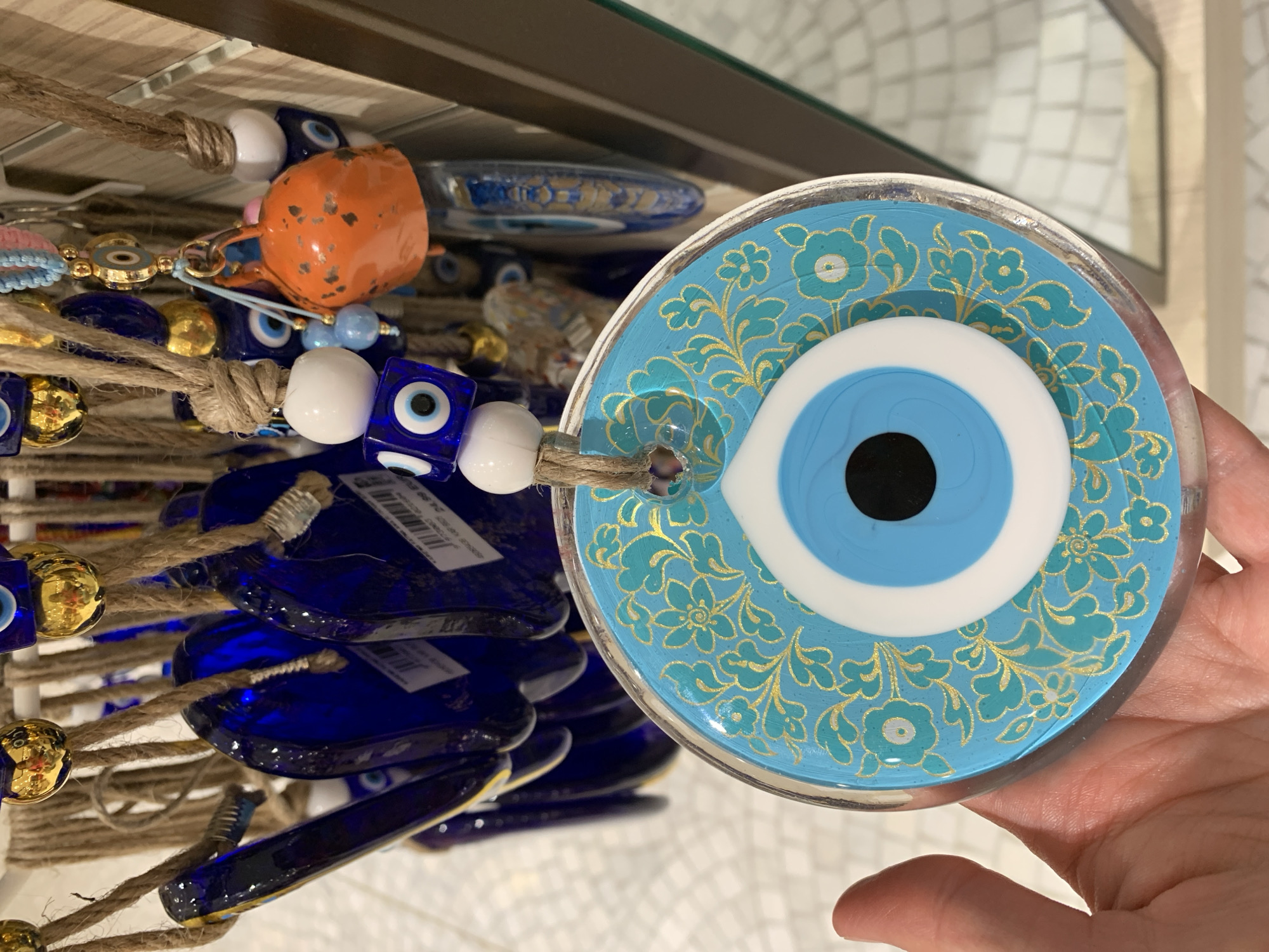 The image shows a close-up of several "evil eye" charms hanging on a display. In the foreground, a person's hand is holding a large, round, blue evil eye charm decorated with gold floral patterns. The charm features a distinct blue and white eye design in the center. Behind this, other blue, tear-drop shaped evil eye charms are visible, as well as cube-shaped blue and white evil eye beads. A small, orange, bell-shaped charm is also present. The charms are suspended by twine. The background suggests a retail environment, with a shelf and other merchandise. The scene appears to be a display of souvenirs or decorative items, possibly in a shop. The context of Aksu, Turkey, implies this is likely a market or tourist shop selling traditional Turkish talismans.