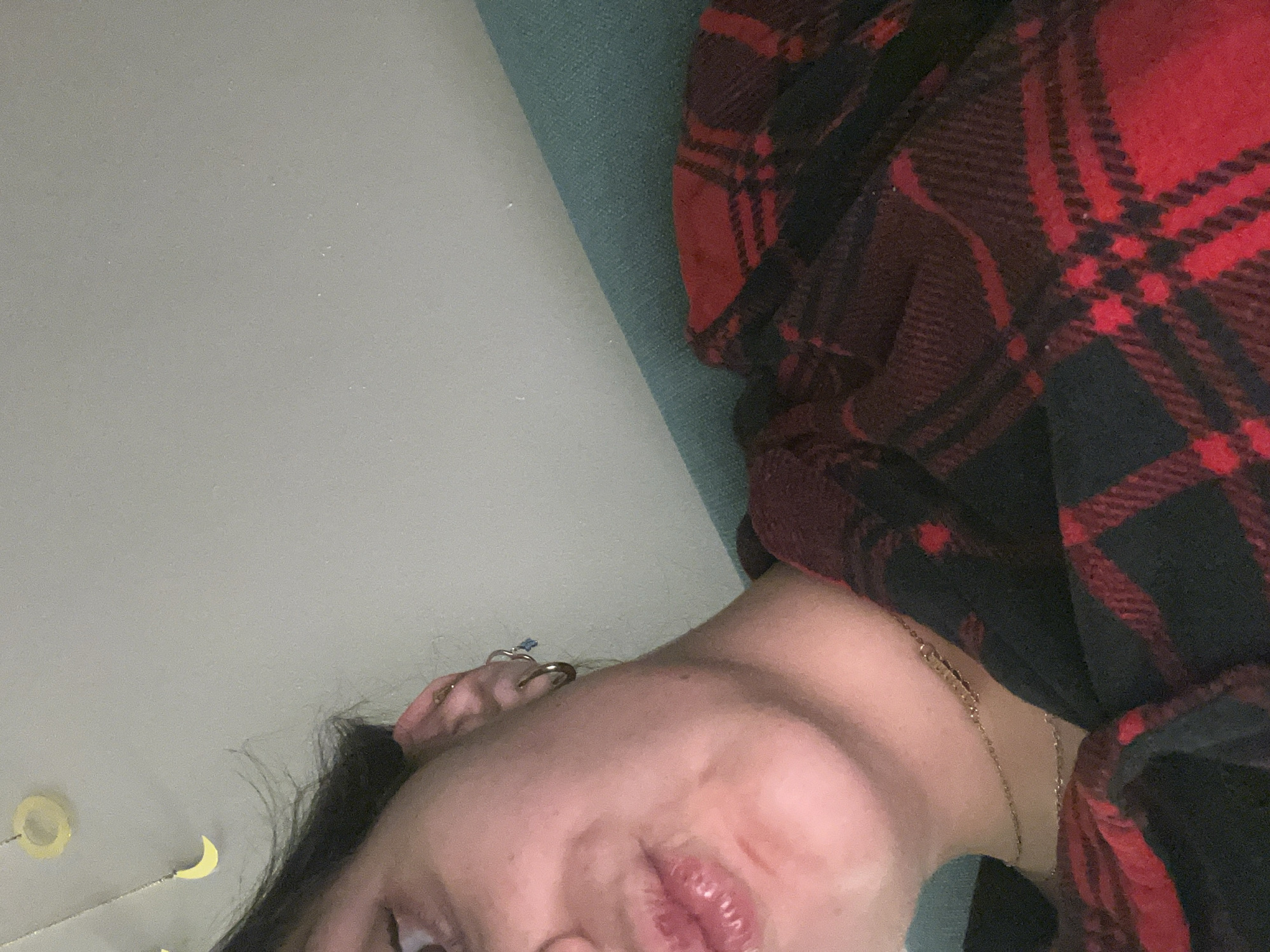 A close-up, angled view of a person. The person appears to be wearing a red and black plaid shirt or blanket, which is draped around their neck and shoulder. A gold necklace is visible around their neck. In the background, there is a neutral-toned wall and a section of what appears to be a blue-green upholstered surface. The location is unknown.