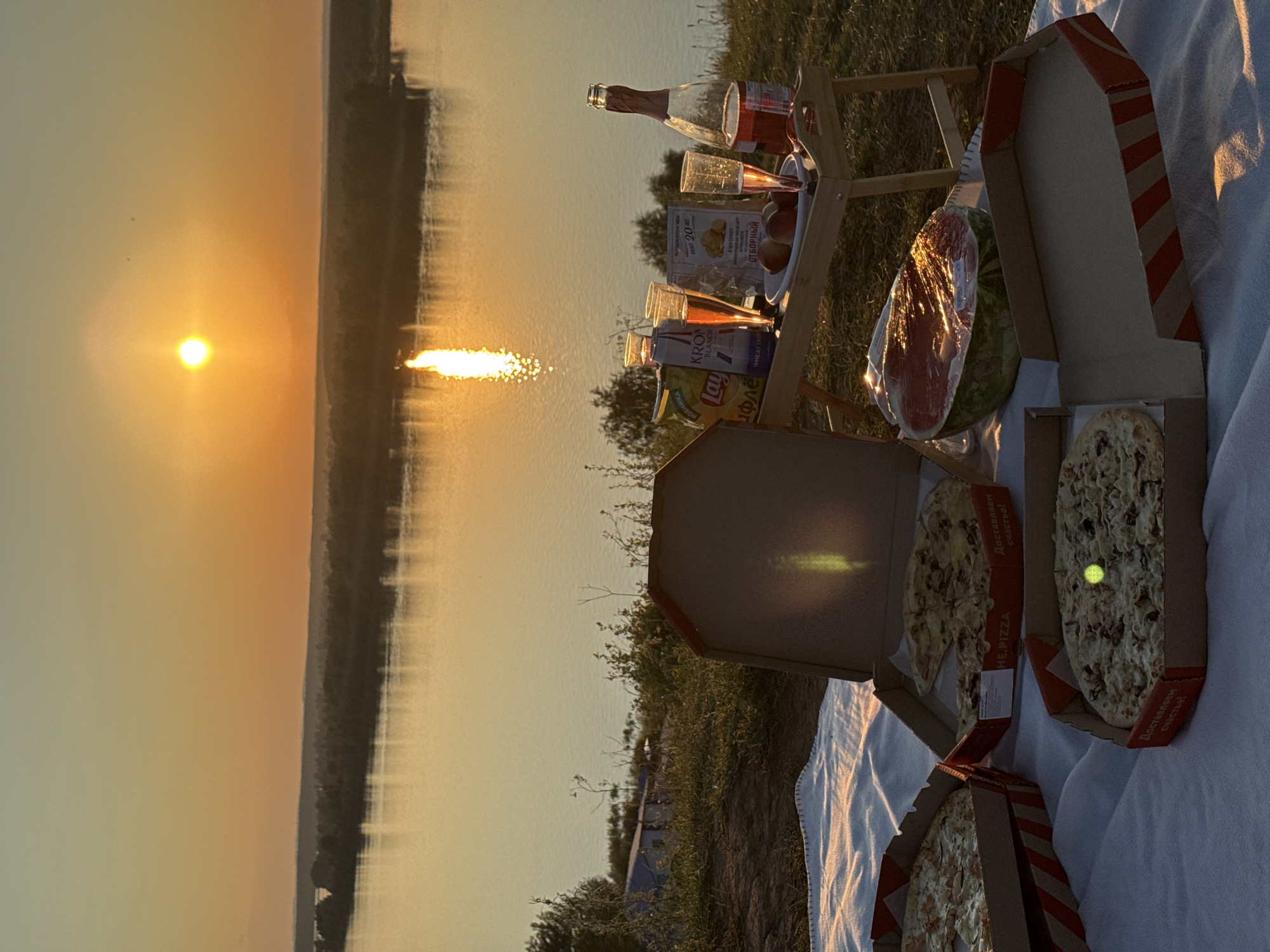 A picnic is set up on a grassy hillside overlooking a wide river at sunset. Three open pizza boxes with pizzas are spread on a white blanket. A half watermelon wrapped in plastic is next to the pizzas. A wooden tray with two glasses of pink beverage, a bottle of pink beverage, a bag of chips, and peaches is placed on the grass. The sun is setting over the horizon, casting a golden glow on the scene. The location is the city of Olkhovka, Russia.