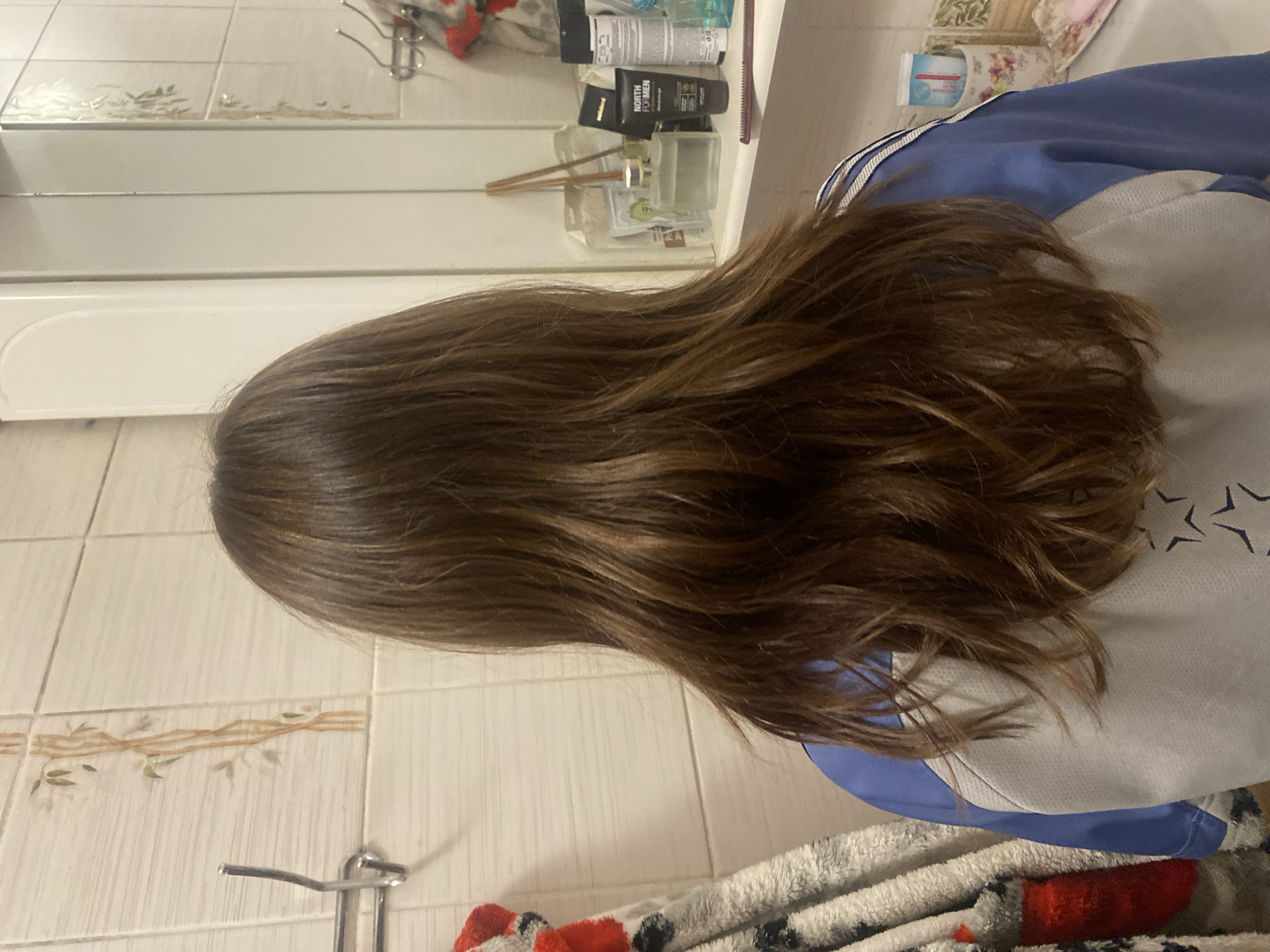 The image shows the back of a person's head with long, brown hair, partially obscuring a tiled wall and a mirror. The person is wearing a blue and white athletic top. In the background, on a shelf in front of the mirror, are various toiletries including a diffuser, bottles, and tubes of products. A towel is hanging on a hook to the left. The scene appears to be in a bathroom. The location is specified as Chornomorsk, Ukraine.