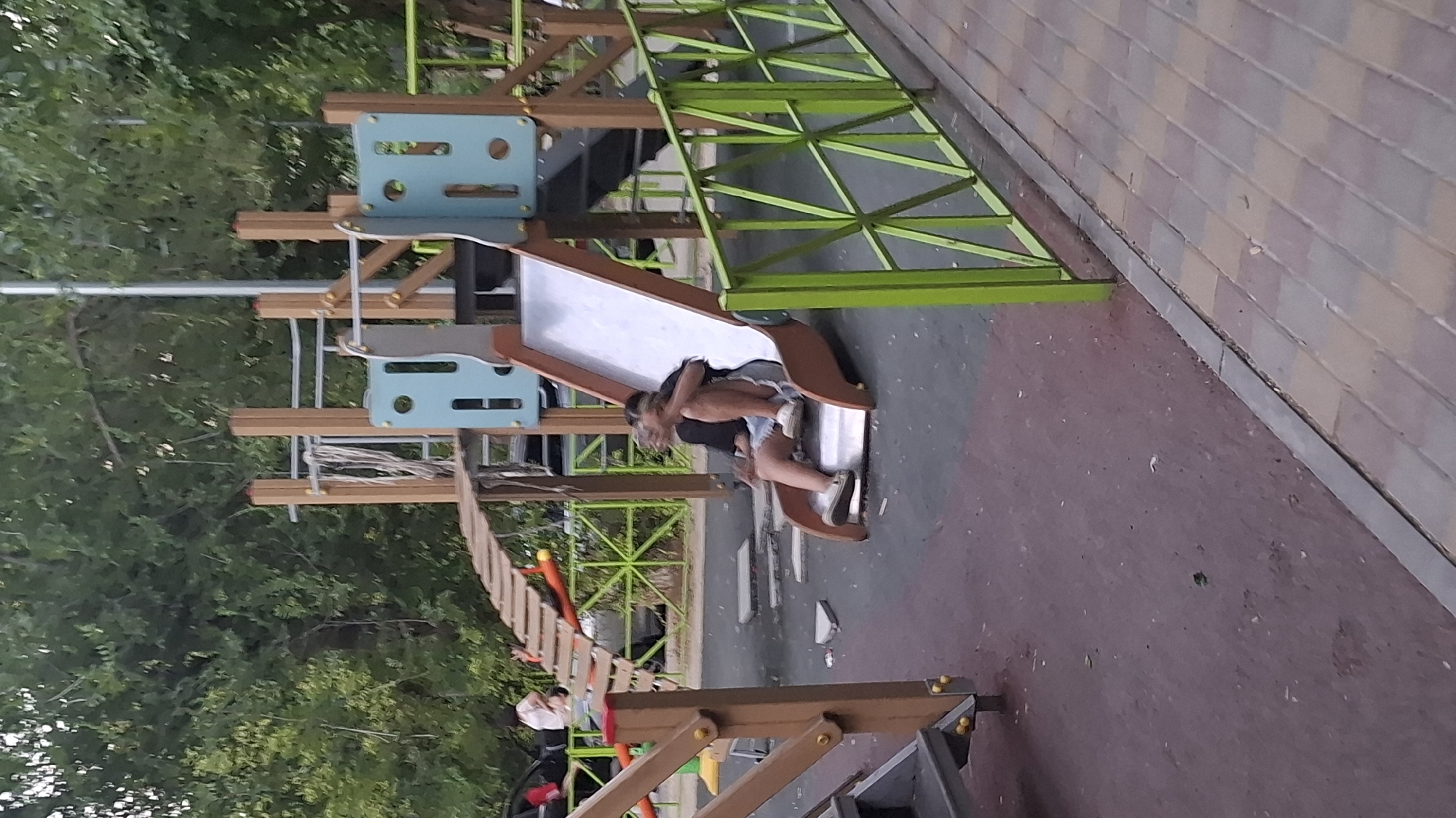 The image shows a playground in the city of Shymkent, Kazakhstan. A young person is sitting on a silver slide, with their legs bent and tucked towards their chest. They are wearing a dark-colored t-shirt, light-colored shorts, and light-colored sneakers. The slide is part of a larger playground structure made of wood and light blue plastic panels. In the background, there are trees and a green metal fence. The ground beneath the slide is a dark, rubberized surface.