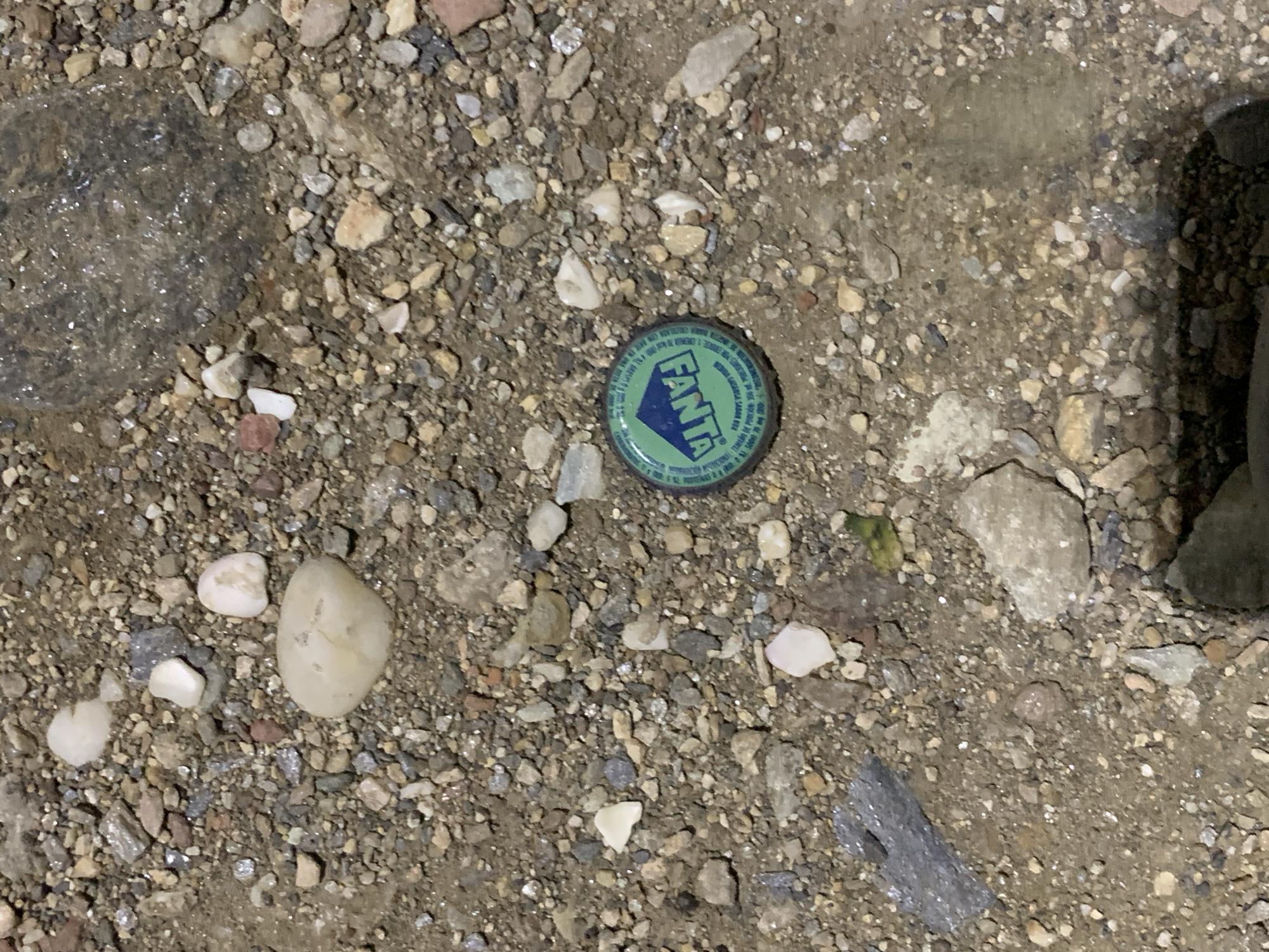 The image displays a ground-level view of an outdoor surface in Santa Ana, Venezuela, predominantly composed of mixed gravel, small stones, and dirt. A single, green Fanta bottle cap, featuring an inverted blue "FANTA" logo and additional white text along its corrugated rim, is positioned in the lower-right quadrant of the frame. The gravel consists of various sizes and shapes of pebbles in hues of white, grey, brown, and reddish-brown, intermingled with fine dirt. Several patches of the ground appear wet and exhibit reflective properties, particularly in the upper-right and central-lower areas. Miscellaneous debris includes a dark, irregularly shaped reflective substance in the upper-right corner and an elongated, grey, reflective object in the mid-left. A small, bright green fragment is also visible immediately to the upper-right of the Fanta bottle cap. No individuals, animals, or active subjects are present within the scene. The lighting suggests artificial illumination, producing distinct reflections on wet surfaces.