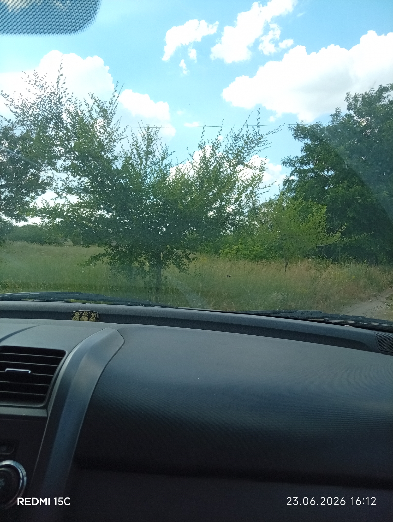 The image is taken from inside a vehicle, looking out through the windshield. The foreground shows the dark dashboard of the car, with a silver vent on the left and a small religious icon depicting three figures on the dashboard. Beyond the car's interior, the scene is outdoors.
The main subjects are trees and vegetation. In the middle ground, there is a field of tall, dry grass, with several trees of varying heights and densities. The trees on the left are fuller and greener, while those on the right are darker and appear to be further away, forming a denser treeline. The background is a bright blue sky with scattered white clouds.
The image appears to have been taken during the daytime, likely in the afternoon, given the lighting and the timestamp visible in the bottom right corner. The weather seems to be clear and sunny, with fluffy white clouds suggesting fair weather.
The visible text in the image consists of a timestamp "23.06.2026 16:12" in the bottom right corner, and "REDMI 15C" in the bottom left corner, likely indicating the device used to capture the photo. The location context provided is Mykolaivka, Ukraine. No specific activity or event is clearly depicted, it appears to be a snapshot of a scenic view from a car journey. There are no discernible emotions conveyed.