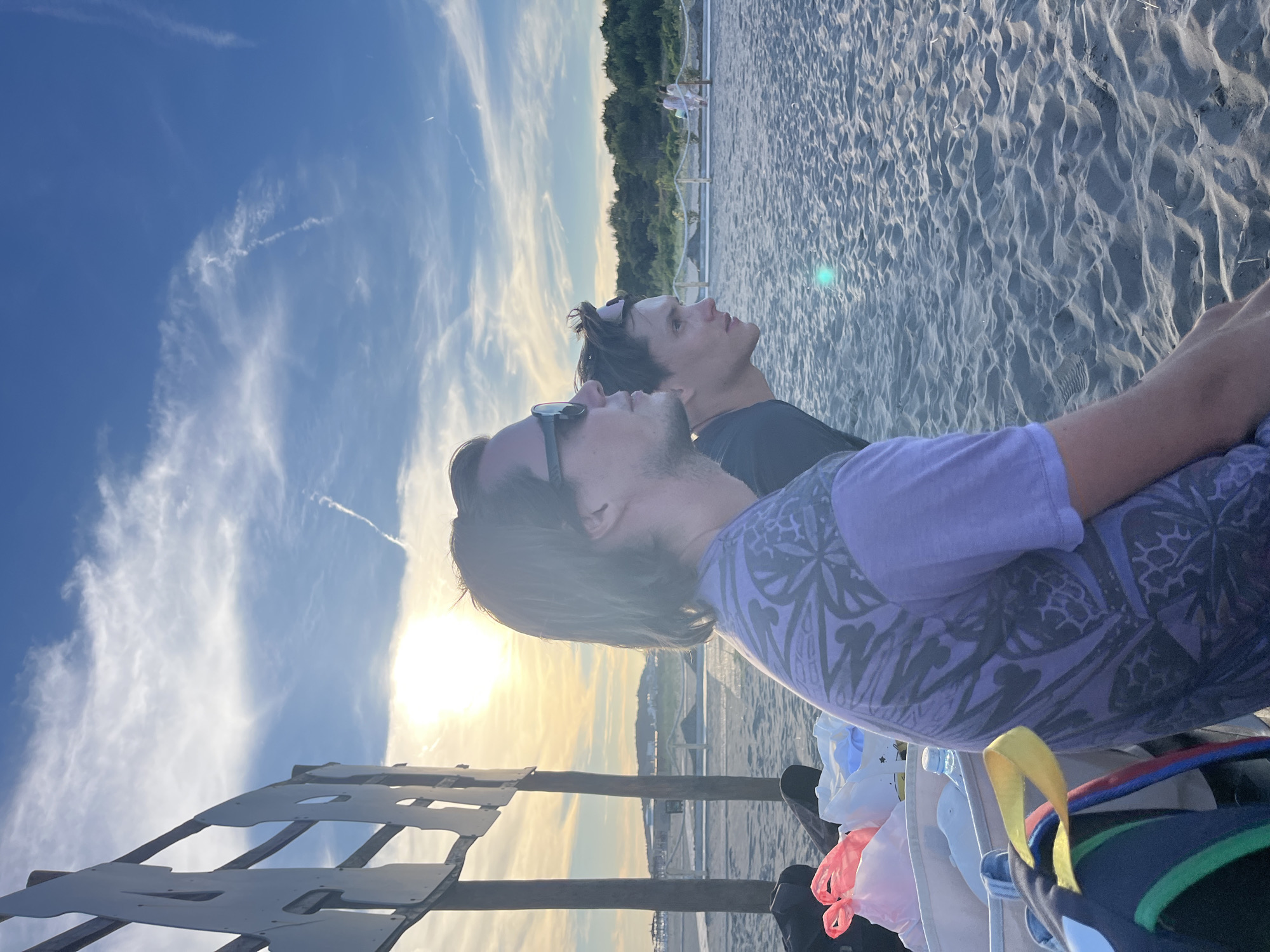 Two individuals are seated on a sandy beach in front of a low wooden structure. The person on the left has long, dark hair and is wearing a purple t-shirt with a patterned design. They are also wearing sunglasses. The person on the right has shorter, darker hair and is looking to their right. They are wearing a black t-shirt and sunglasses perched on their head. In the foreground, to the left of the individuals, are several bags, including a white tote bag with some items inside, a pink plastic bag, and a backpack with colorful straps. The background shows a beach area with more sand and a distant treeline. The sky is blue with scattered clouds and the sun is visible, casting a warm glow. The scene appears to be set in the city of Pistula, Montenegro.