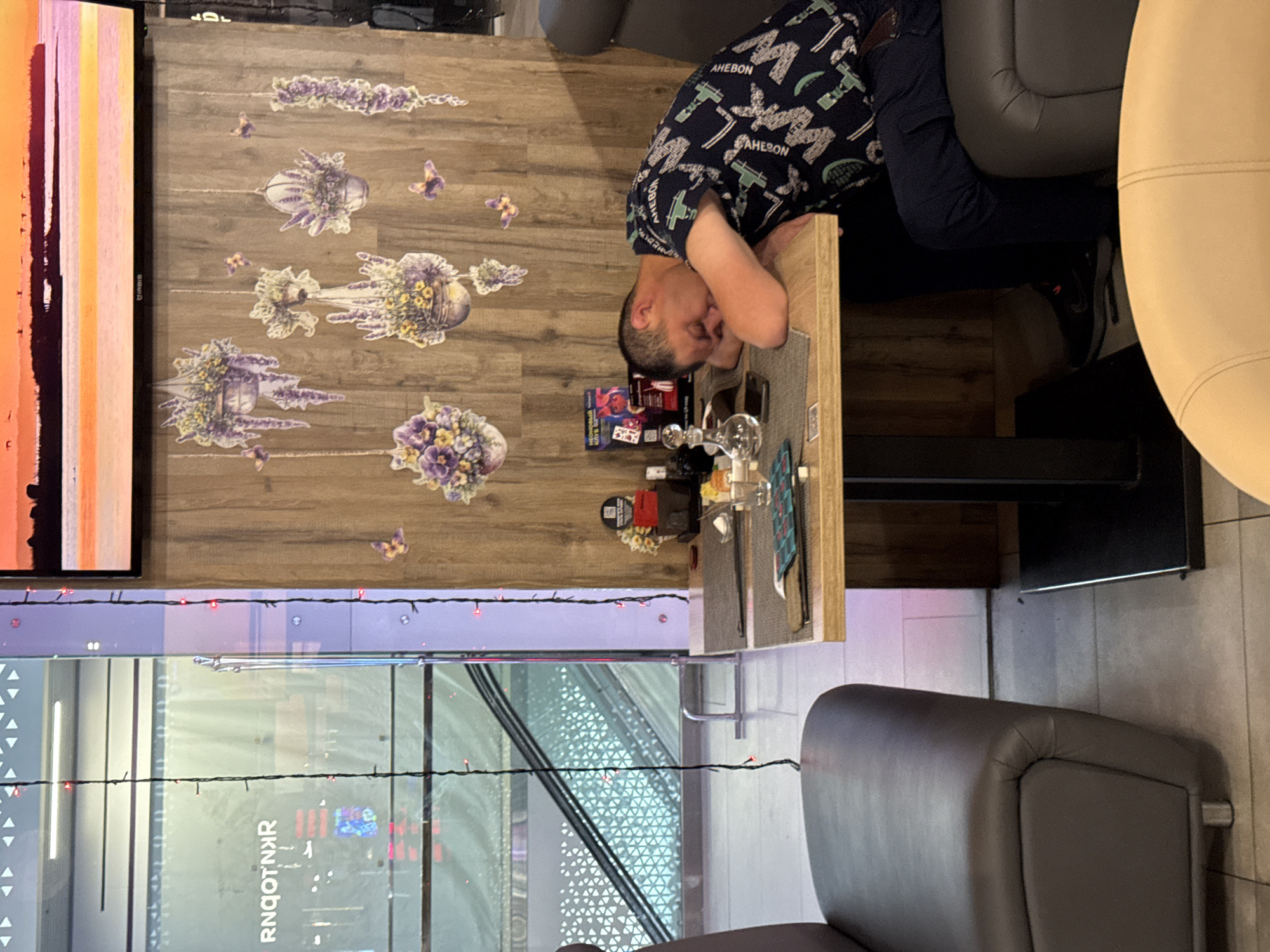 A man in a dark patterned shirt and dark pants sits at a table with floral decorations in what appears to be a restaurant or cafe in Domodedovo, Russia. The man has his head down on the table and may be sleeping. On the table are glasses and other items. Next to the table is a grey chair. A partially visible TV screen shows colorful stripes. Below the table is a view of a city street with signs in Russian. The scene appears to be indoors, possibly inside an establishment overlooking the street.